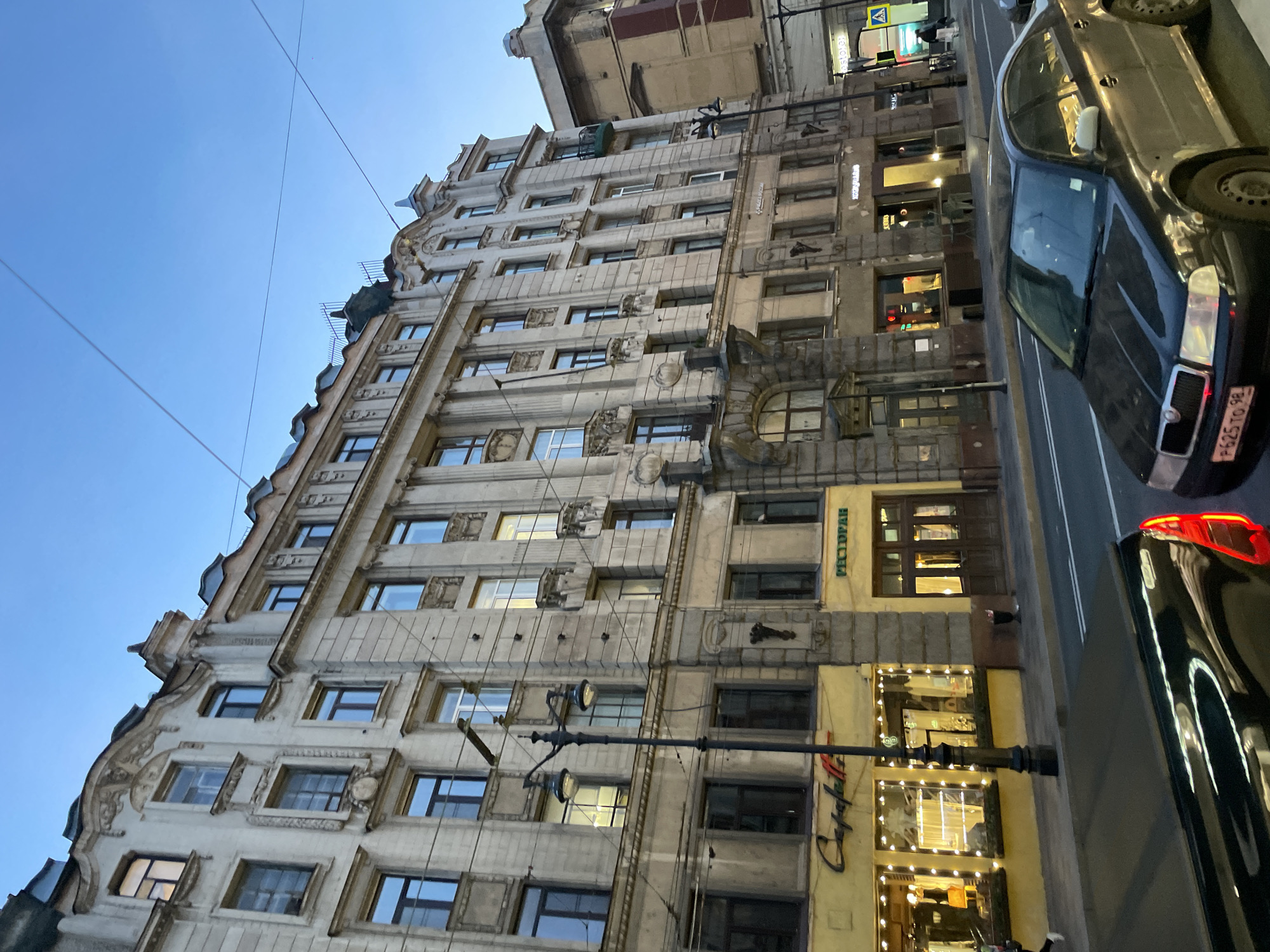 This image shows a tall, ornate building with many windows in Saint Petersburg, Russia. The building is predominantly light-colored with detailed architectural features around the windows and along the roofline. On the right side of the building, at street level, there is a storefront with signage in Russian; one sign reads "Cafefella." The street is to the right of the building, with several cars partially visible. The sky above is a clear, light blue. Utility poles and wires are also visible in the sky.