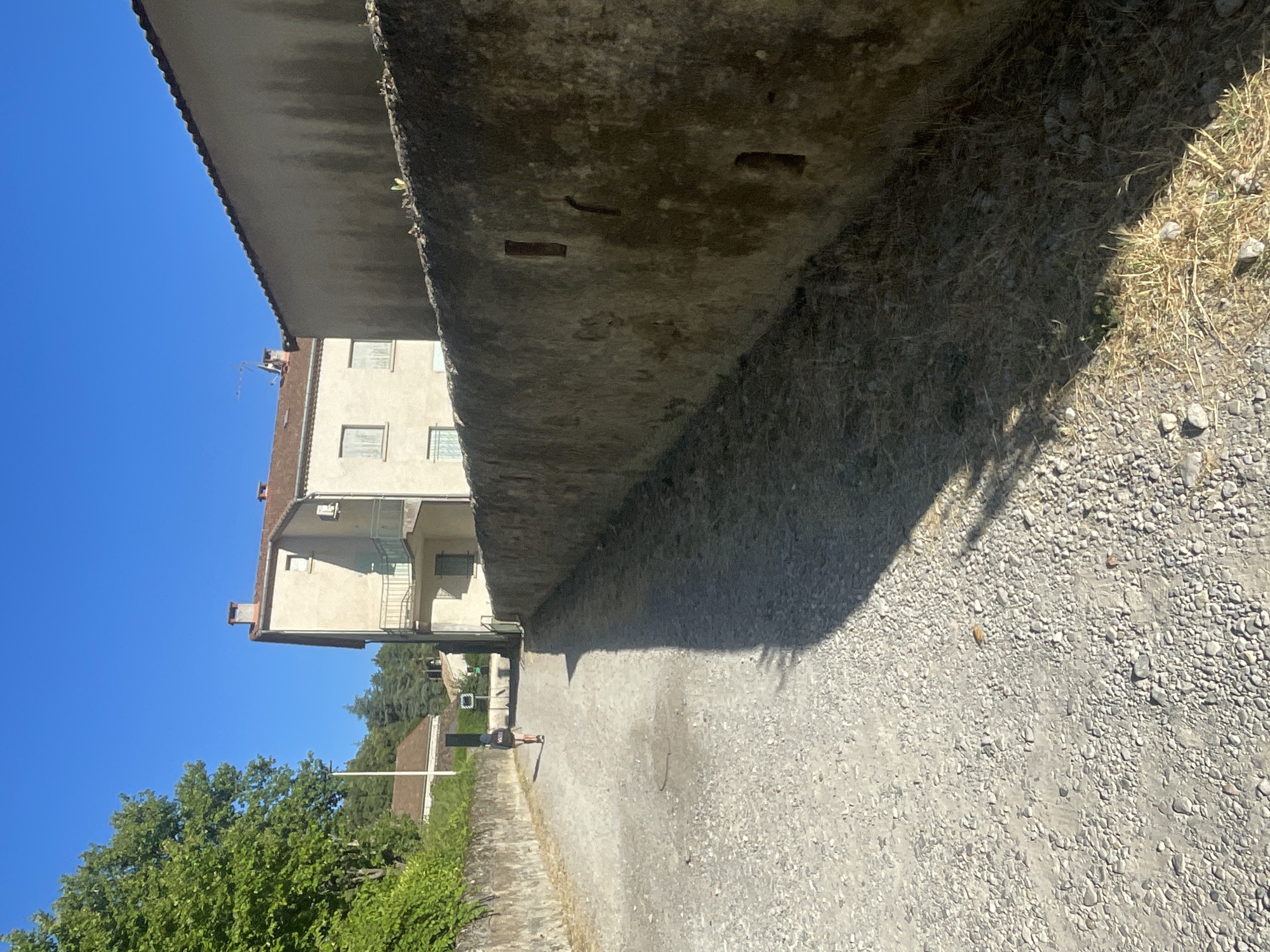A full shot shows a person walking away from the camera down a gravel path lined with stone walls. The person is wearing a dark backpack and light-colored shorts. The path leads to buildings in the distance, including a multi-story building with a red roof and a light-colored facade. To the right of the path is a tall, textured stone wall with several small, recessed openings. The scene is set under a clear blue sky with bright sunlight casting a long shadow from the wall onto the path. Lush green trees are visible on the left side of the image.