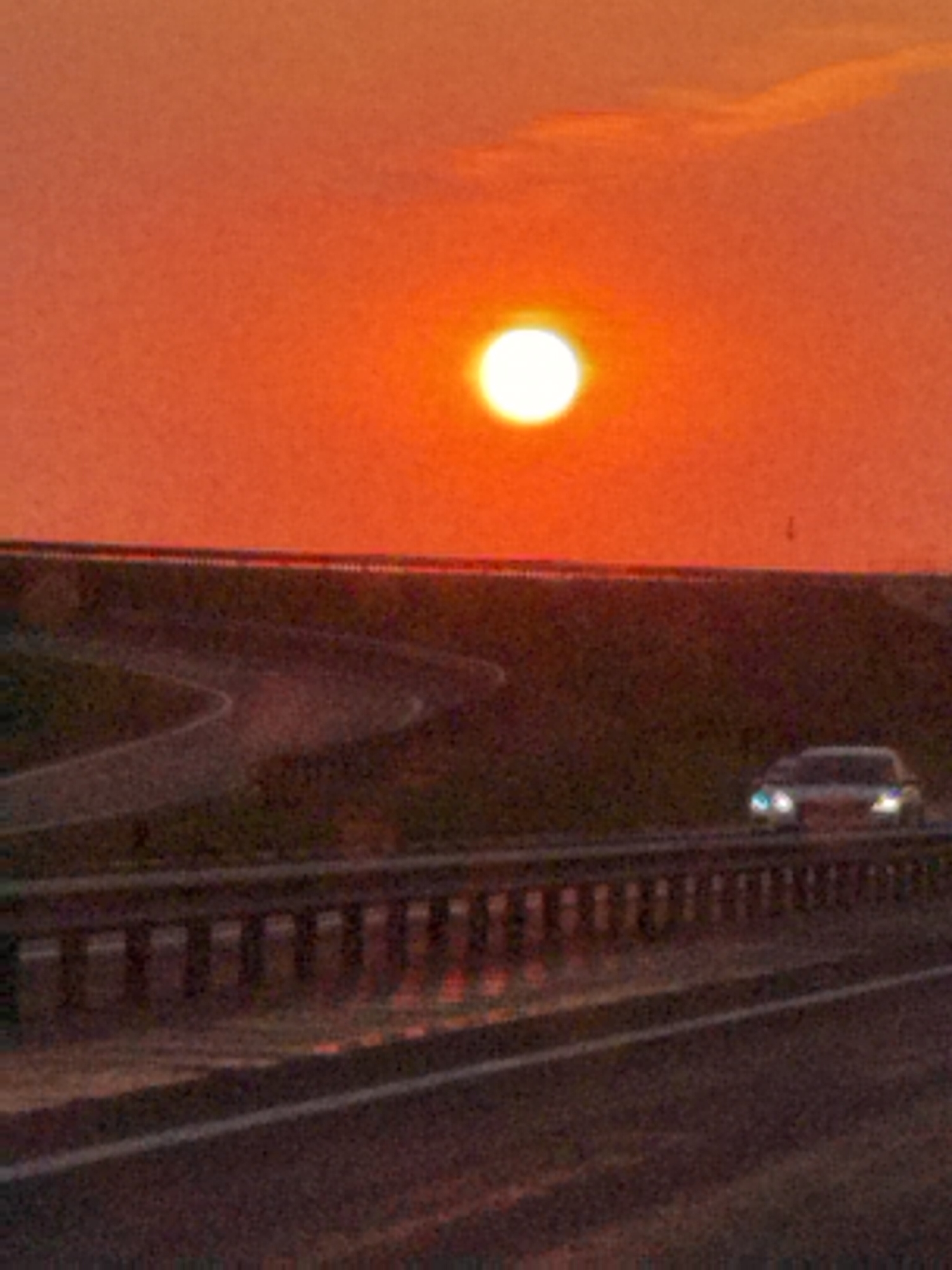 The image shows a sunset over a highway in Абухаўскі сельскі Савет, Belarus. The sun, a bright orb, is low on the horizon, casting a warm orange glow across the sky and clouds. A highway with a guardrail is visible in the foreground, with a car driving on it. The landscape appears to be rural, with trees and fields in the background. The overall mood of the image is peaceful and serene.