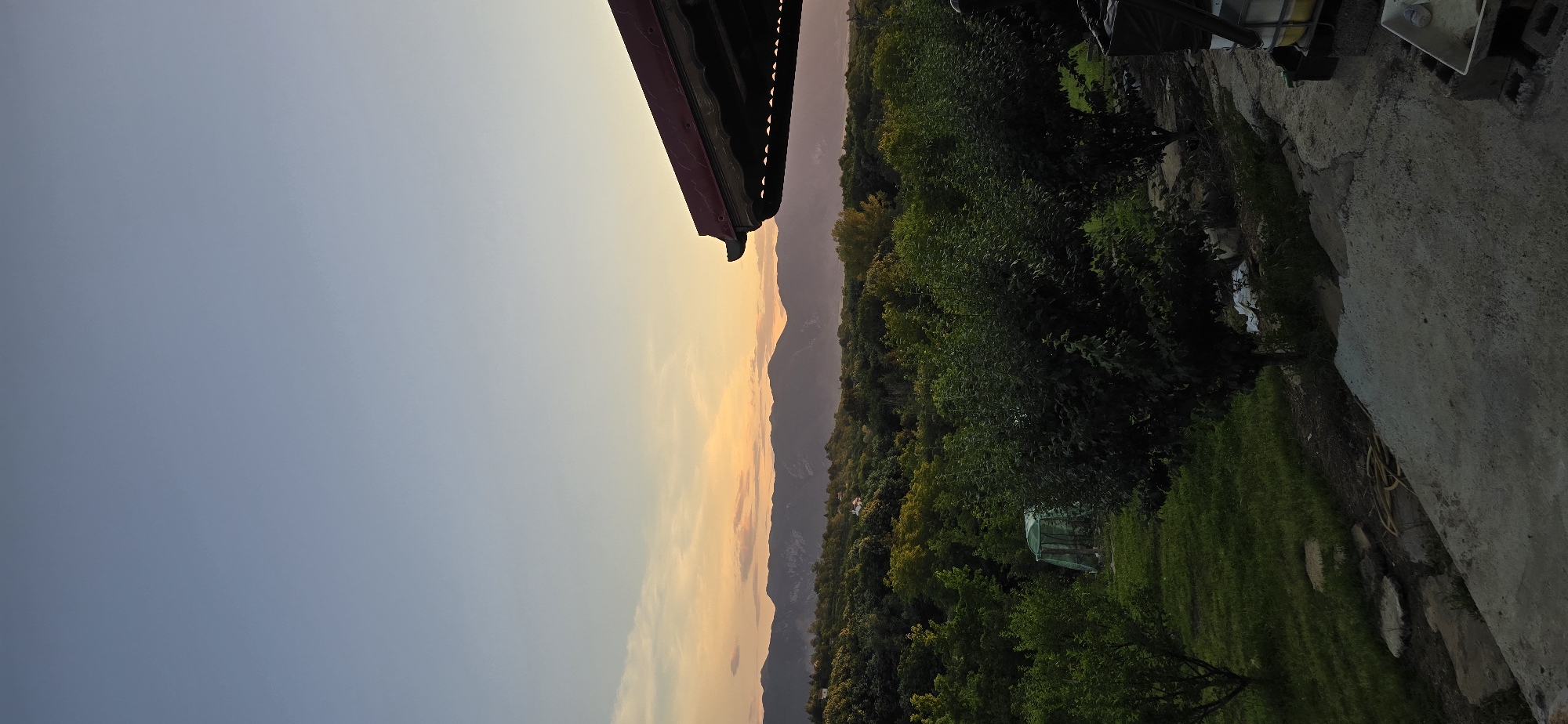 The image shows a panoramic view of a rural landscape at dusk. In the foreground, a concrete patio with some scattered stones and a yellow hose is visible. To the right, the corner of a red-tiled roof extends into the frame, supported by dark wooden beams and featuring decorative cutouts. Beyond the patio, a lush green lawn is dotted with trees and a small green greenhouse. In the middle ground, a dense forest of varying shades of green stretches across rolling hills. In the background, a range of majestic mountains forms a silhouette against the horizon, bathed in the warm glow of the setting sun. The sky transitions from a pale blue at the top to a soft orange and pink hue near the mountains, with faint wispy clouds. There are no people visible in the image. The location is described as Unknown, Unknown.