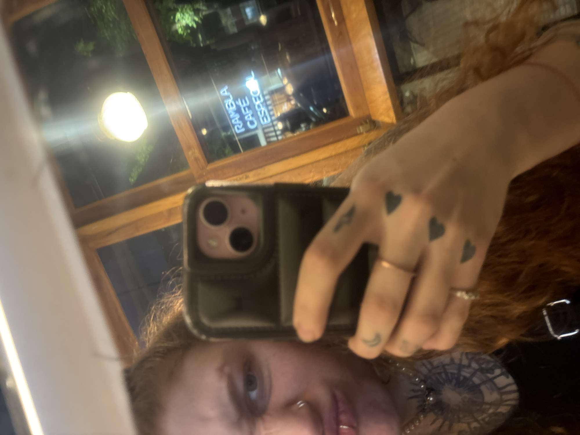 A selfie taken in Buenos Aires, Argentina. The individual is holding a phone, obscuring their face, but visible details include red hair, facial piercings, and tattoos on their hand (hearts and a crescent moon). The reflection in the window behind them shows a light fixture and the signage for "RAMBLA CAFÉ ESPECIAL," indicating a cafe environment. The image is slightly blurry and taken at an angle.