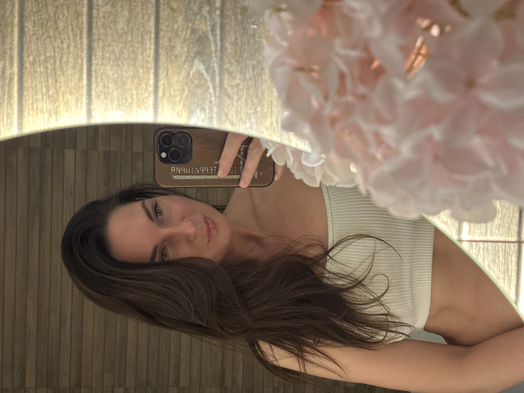 A brunette woman is taking a selfie in a mirror, with the reflection of a wall with vertical panels visible in the background. She is holding her phone, which has a wooden case with writing on it. She is wearing a light-colored ribbed top, and artificial flowers are in the upper right corner of the mirror's reflection. The location is Chaikovsky, Russia.