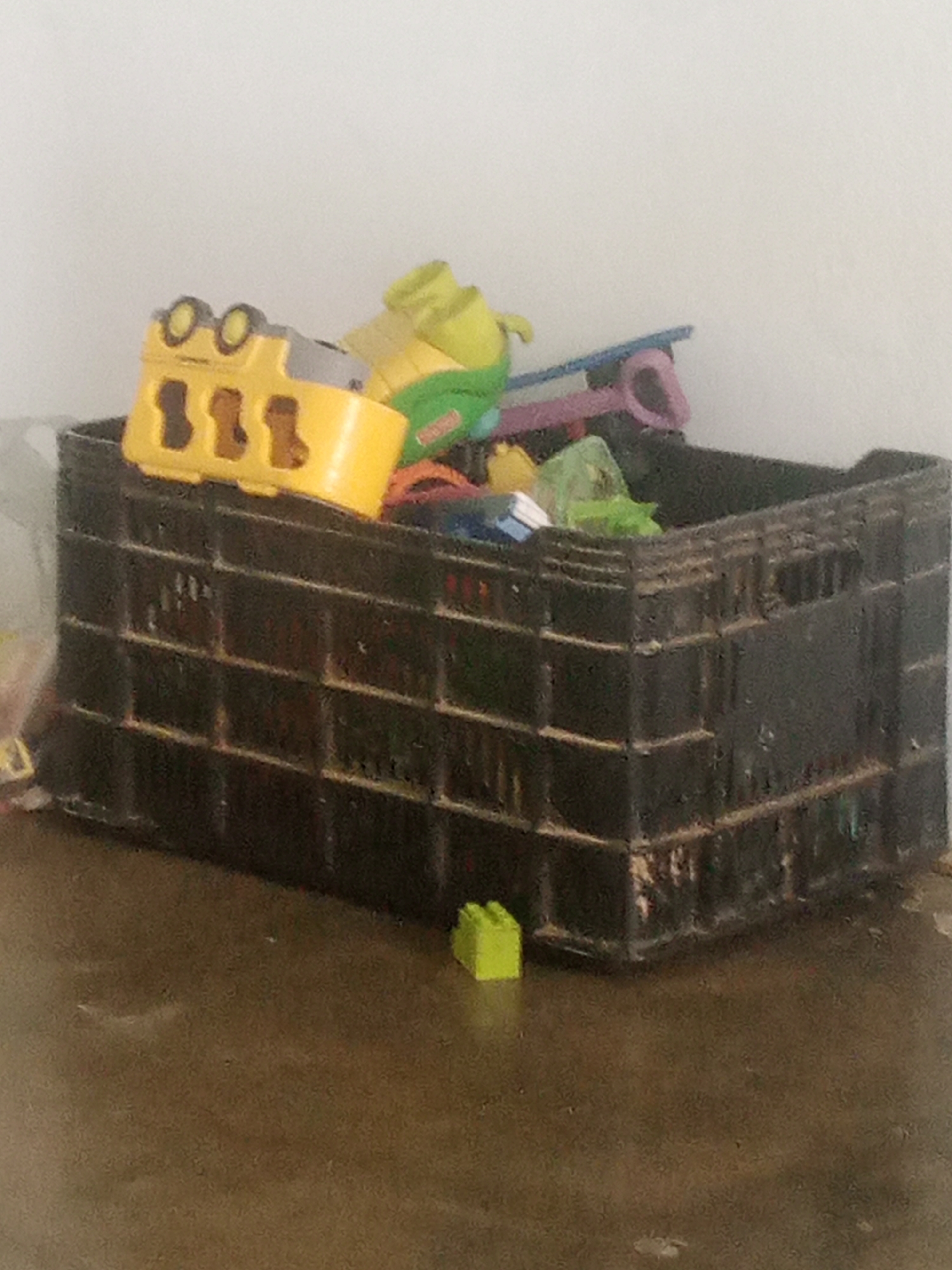 A black plastic crate filled with toys sits on a wet, brown surface. The toys include a yellow bus-like vehicle with a gray roof and black wheels, a green dinosaur-shaped toy, and other colorful plastic pieces, some appearing to be building blocks. A single green building block rests on the wet surface to the right of the crate. The background is a blurred white or light-colored wall. The scene appears to be outdoors, possibly in La Tagua, Venezuela, given the wet environment and the general appearance. There are no people or animals visible in the image.