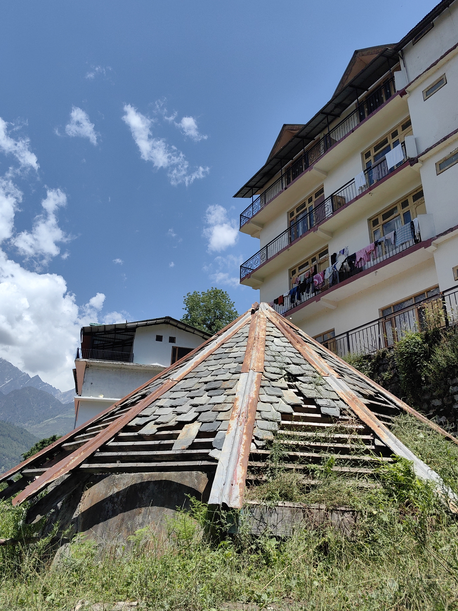 The image depicts a multi-layered scene in Vashisht, India, under a clear blue sky with scattered white cumulus clouds.
In the immediate foreground, there is a partially collapsed, roughly conical or pyramidal roof structure. This structure is composed of unevenly laid, dark grey slate tiles, many of which are missing or broken, exposing light-colored wooden laths underneath. Rusted corrugated metal sheets or supports run along the edges of the structure. The base of this roof is surrounded by dense, green vegetation, including grass and weeds, which also grows through gaps in the roof structure. Beneath the roof, a dark, curved, weathered wall or foundation is partially visible.
In the midground, to the right, stands a multi-story building painted in a light cream or off-white color. It features at least three visible floors, each equipped with a balcony. The balconies have black metal railings with decorative patterns and a reddish-brown trim along their bases. Multiple wooden-framed windows are visible across the building's facade. Clothes of various colors and types, including shirts, trousers, and towels, are hanging to dry on the railings of the lower two visible balconies. The building's roof has a dark, possibly corrugated metal, overhang. To the left of this main building, a smaller, white-painted building with a dark roof and a small balcony is partially visible, situated slightly behind the foreground structure. More green foliage and bushes are visible on the terrain between the foreground roof and the main building.
In the background, on the far left, a range of rugged mountains with sharp, rocky peaks is visible under the blue sky. Some lighter patches on the mountain peaks suggest snow or barren rock.