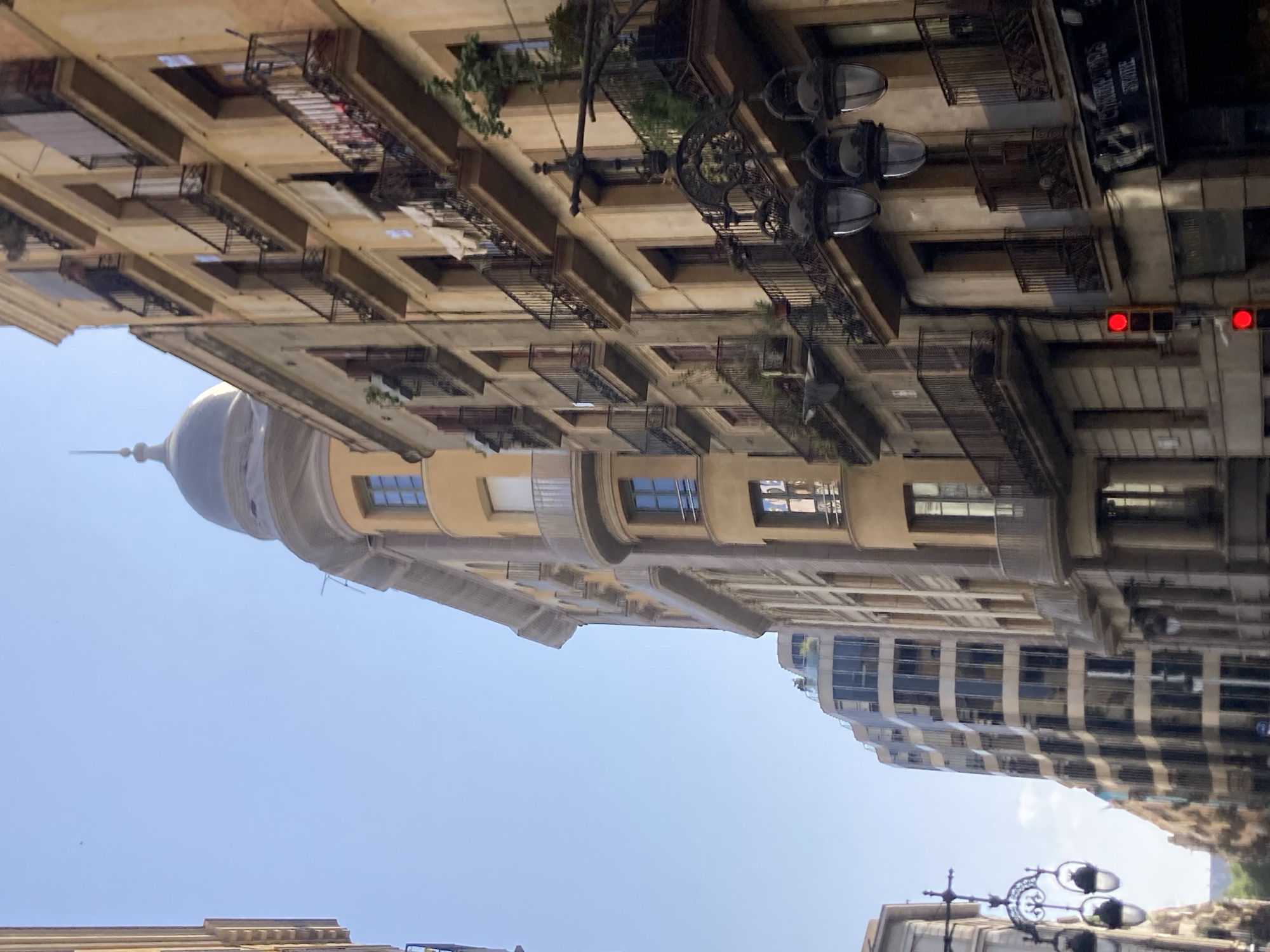 The image shows a street scene in Barcelona, Spain, with a clear blue sky. On the right side of the frame, there are several tall, multi-story buildings with facades of ochre and beige tones. These buildings feature numerous balconies with wrought-iron railings, some of which have plants and laundry hanging on them. The architectural style suggests a historic European city.
A prominent feature on the left side of the image is a modern building with a curved, glass facade, reflecting the sky and surrounding structures. Further down the street, more buildings can be seen, some with ornate lampposts. A traffic light with red lights illuminated is visible in the lower right quadrant of the image, indicating that traffic is stopped. The perspective of the image is looking upwards, emphasizing the height of the buildings. There are no people clearly visible in the image. The overall impression is one of an urban environment bathed in sunlight.