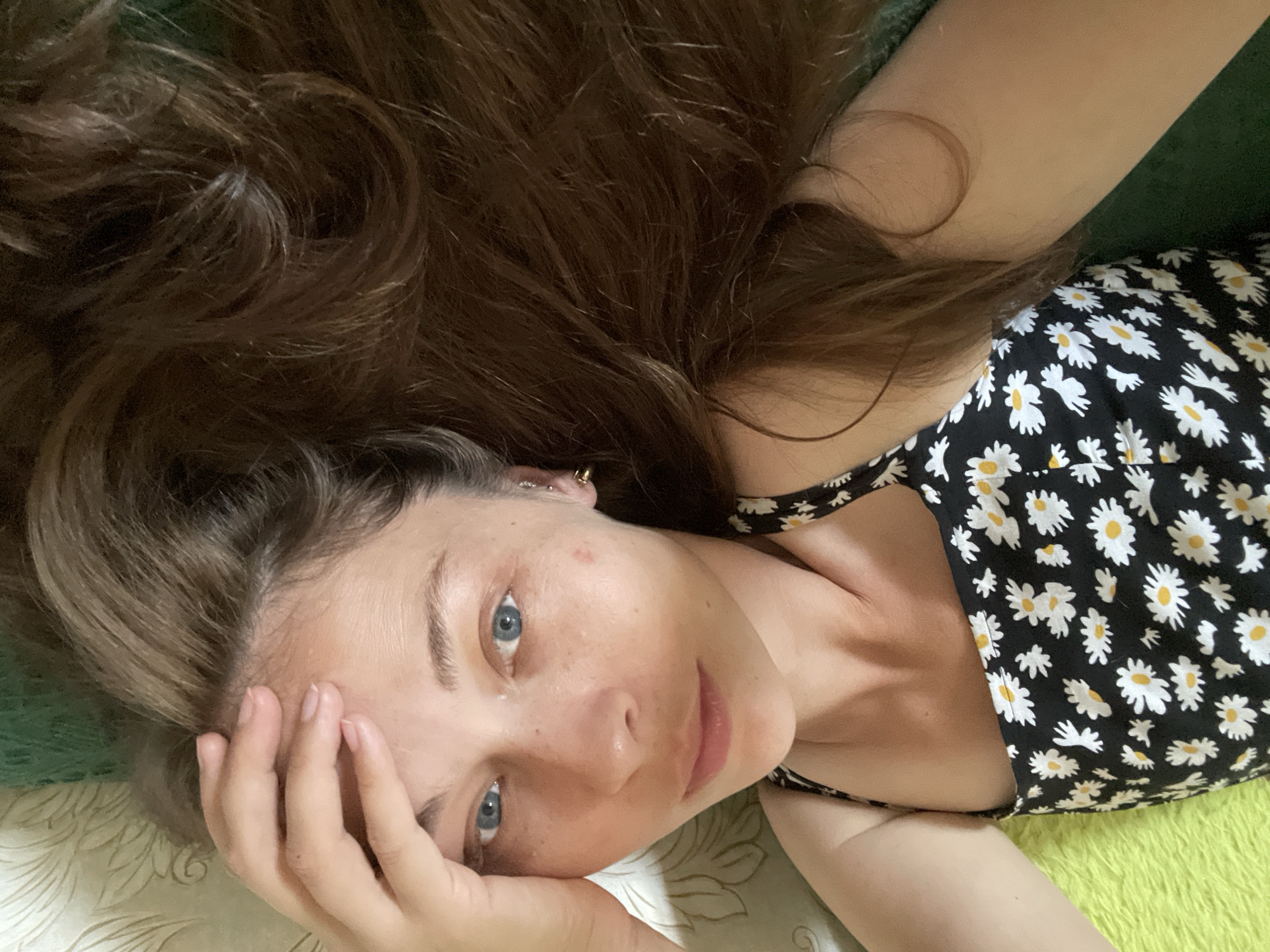 A young woman with long, brown hair is lying on her side, looking directly at the camera. She has striking blue eyes and fair skin, with a few freckles visible on her cheeks and nose. Her left hand is resting on her forehead, and she appears to be relaxed or contemplative. She is wearing a black dress adorned with a pattern of white daisies with yellow centers. The background is out of focus, but appears to be a textured surface in muted tones, possibly a bed or sofa. The lighting suggests it could be daytime, indoors. There is no visible text in the image.