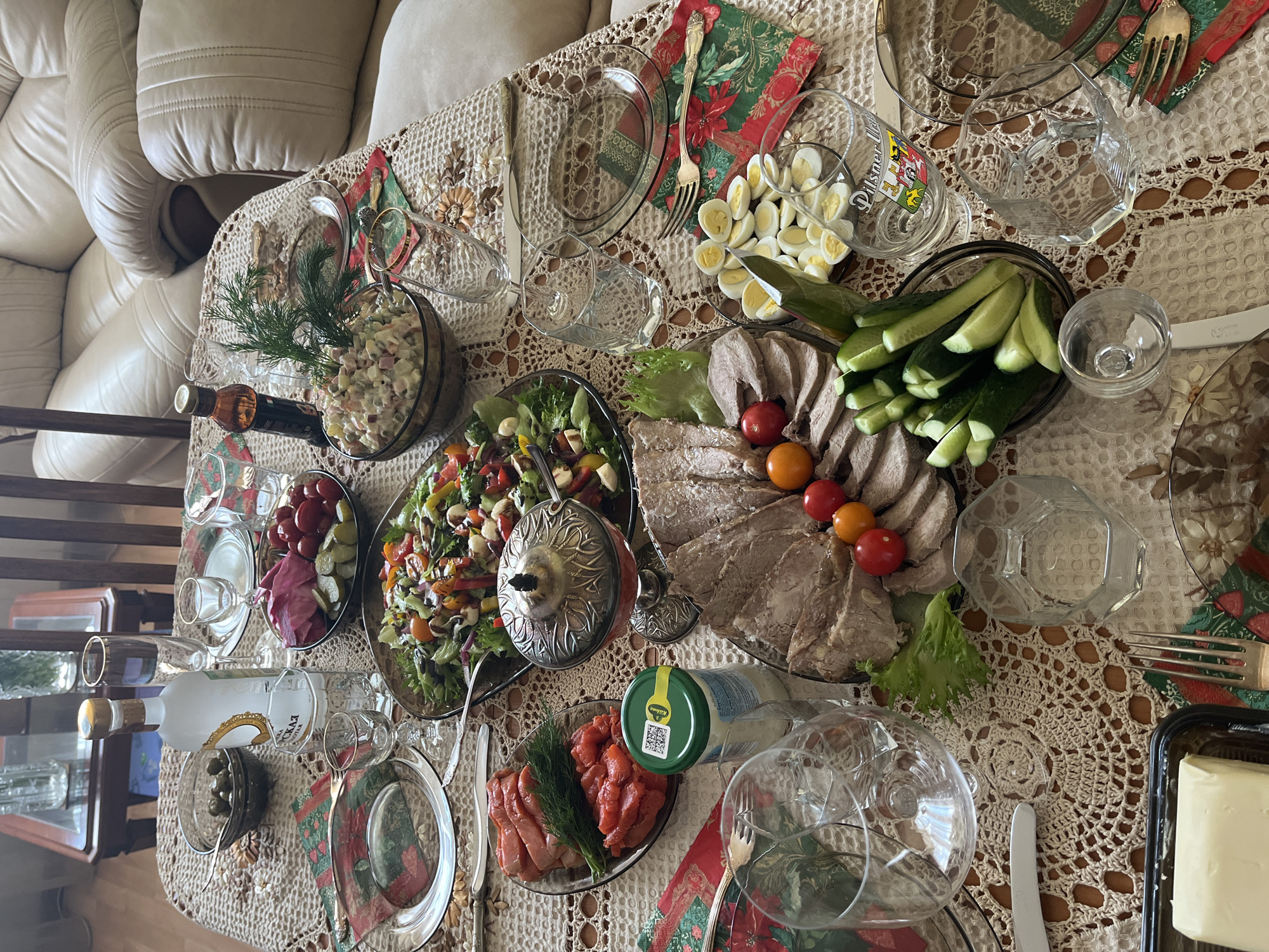 The image shows a table laden with food and drinks, set for a meal. The table is covered with a lace tablecloth. Dishes include Olivier salad, a mixed salad, sliced meat with tomatoes and cucumbers, sliced boiled eggs, sliced salmon, olives, and butter. Alcoholic beverages are present, including vodka and a bottle resembling brandy or whiskey. Silverware is arranged around the plates. Christmas-themed napkins are visible. The setting suggests a festive occasion, possibly a celebration in a home. It is difficult to confirm that the location is in Moscow, Russia without additional clues, though some of the food and drink items, like Olivier salad and vodka, are commonly associated with Russian cuisine and celebrations.