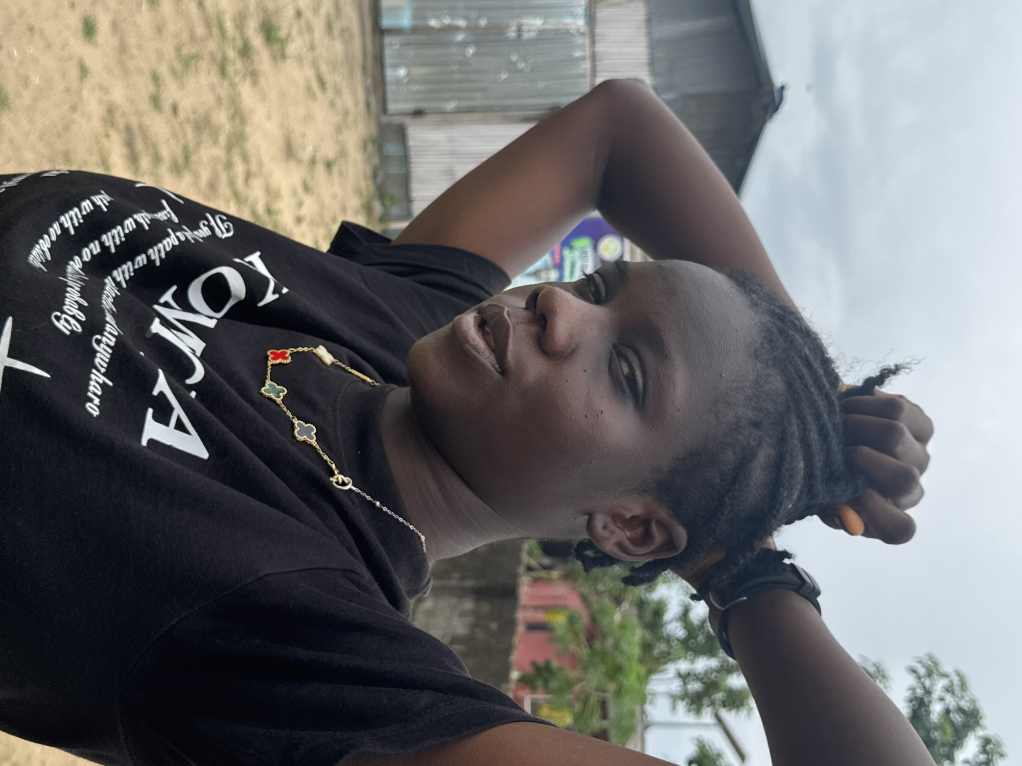 An individual is positioned outdoors with their arms raised above their head, possibly resting behind their head. They are wearing a black T-shirt with white lettering and a necklace featuring a clover design. A watch is visible on the left wrist. The background includes a corrugated metal structure, greenery, and a cloudy sky. A patch of sandy ground is visible in the foreground. The location is identified as Badagry, Nigeria.
