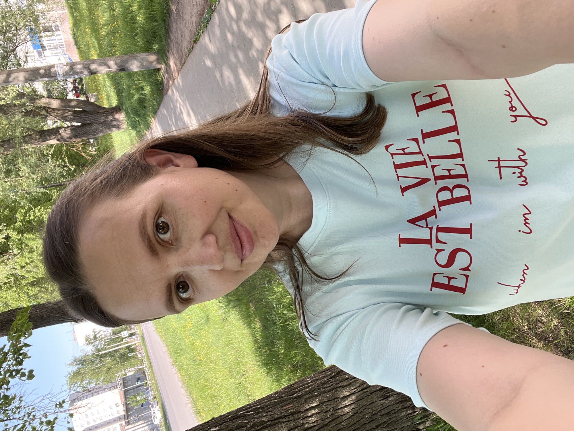A woman is taking a selfie in what appears to be a park or green space. She is wearing a light blue t-shirt with red writing in French, "La Vie Est Belle" and "when I'm with you". The photo is taken from a slightly elevated angle.
The background shows green grass, trees, a sidewalk or path, and a road. In the distance, there are buildings visible.
Based on the buildings visible in the background, it is highly probable that the location is Chaikovsky, Russia. The specific park or green space is not discernible from the image.