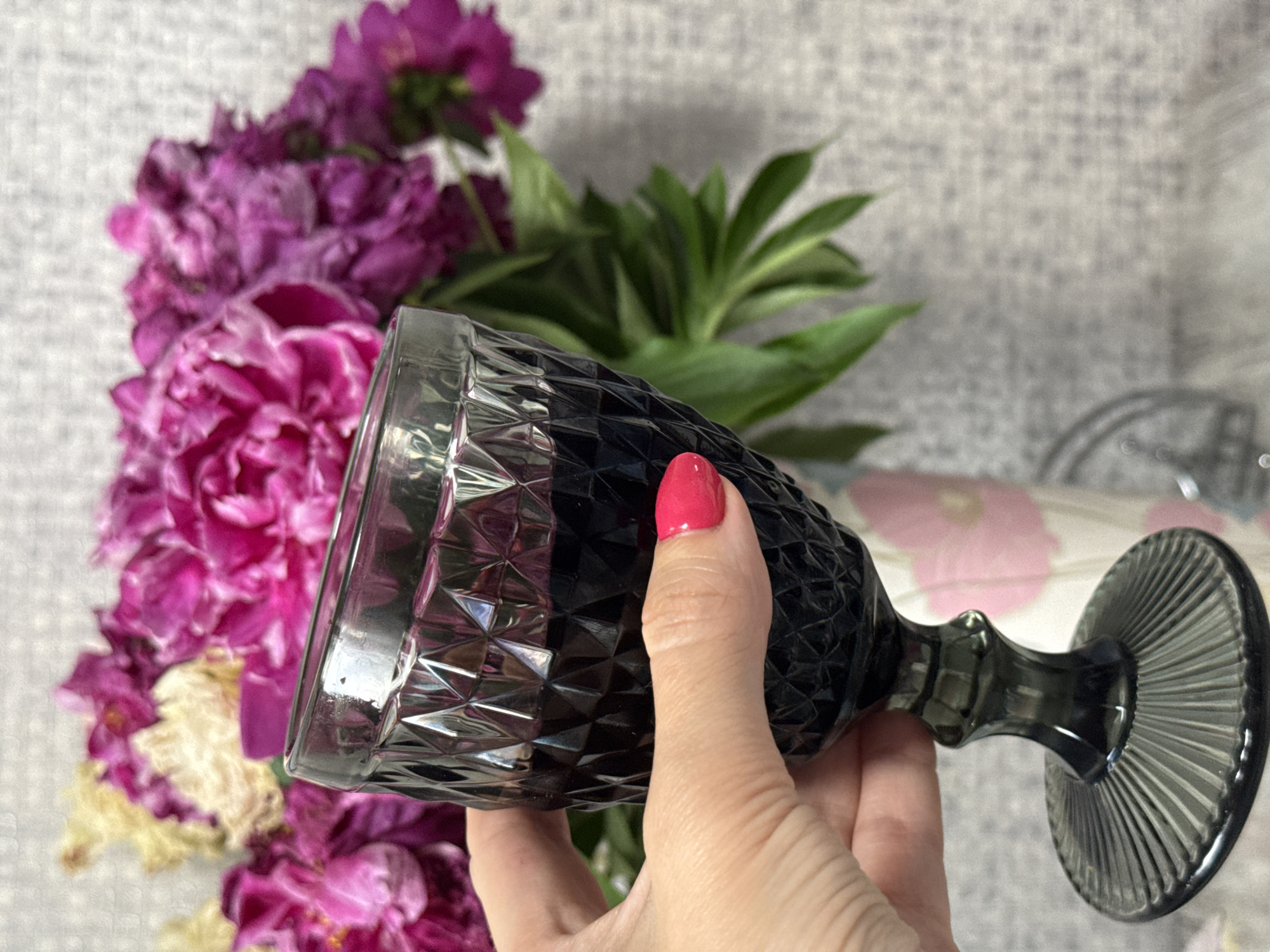A hand with bright pink nail polish is holding a stemmed, dark gray glass filled with a dark liquid. The glass has a diamond-cut pattern on its bowl and a fluted base. In the background, out of focus, is a bouquet of pink and white flowers, possibly peonies, in a vase. The vase appears to have a floral pattern on it. The overall scene is domestic, with a textured gray wall in the background. There is no indication of the location being Moscow, Russia, or any specific city.