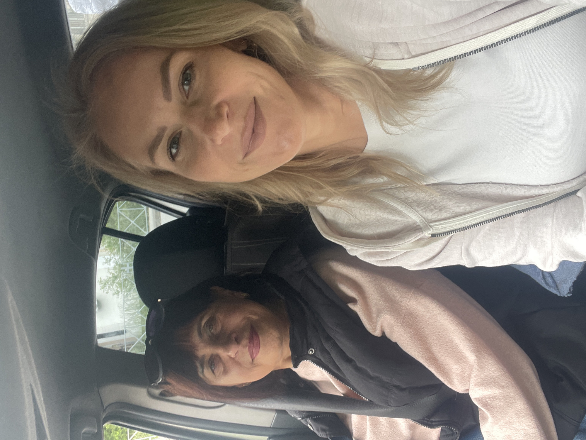 Two women are inside a car. The woman at the top has blonde hair and wears a white shirt and a light pink zipped jacket. The woman at the bottom has short brown hair and wears a light pink shirt and a black vest. Sunglasses are on top of her head. The car is likely traveling in Sochi, Russia. Trees and buildings are visible through the car windows.