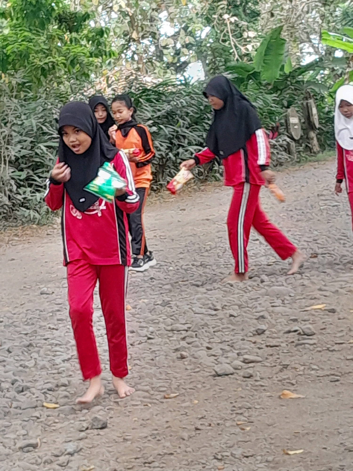 In an outdoor setting with a dirt and gravel path, several young girls are walking. The girl in the foreground on the left is wearing a black hijab and a red tracksuit with white stripes on the sleeves and legs. She is barefoot and holding a green and white snack packet. To her right, another girl is wearing an orange and black tracksuit and is barefoot. Behind them, two other girls are also visible, one in a black hijab and a red and white tracksuit, and another in a white hijab and a red tracksuit, also barefoot. The background consists of lush green foliage and trees. The overall scene suggests a casual outdoor activity, possibly a school outing or a walk.