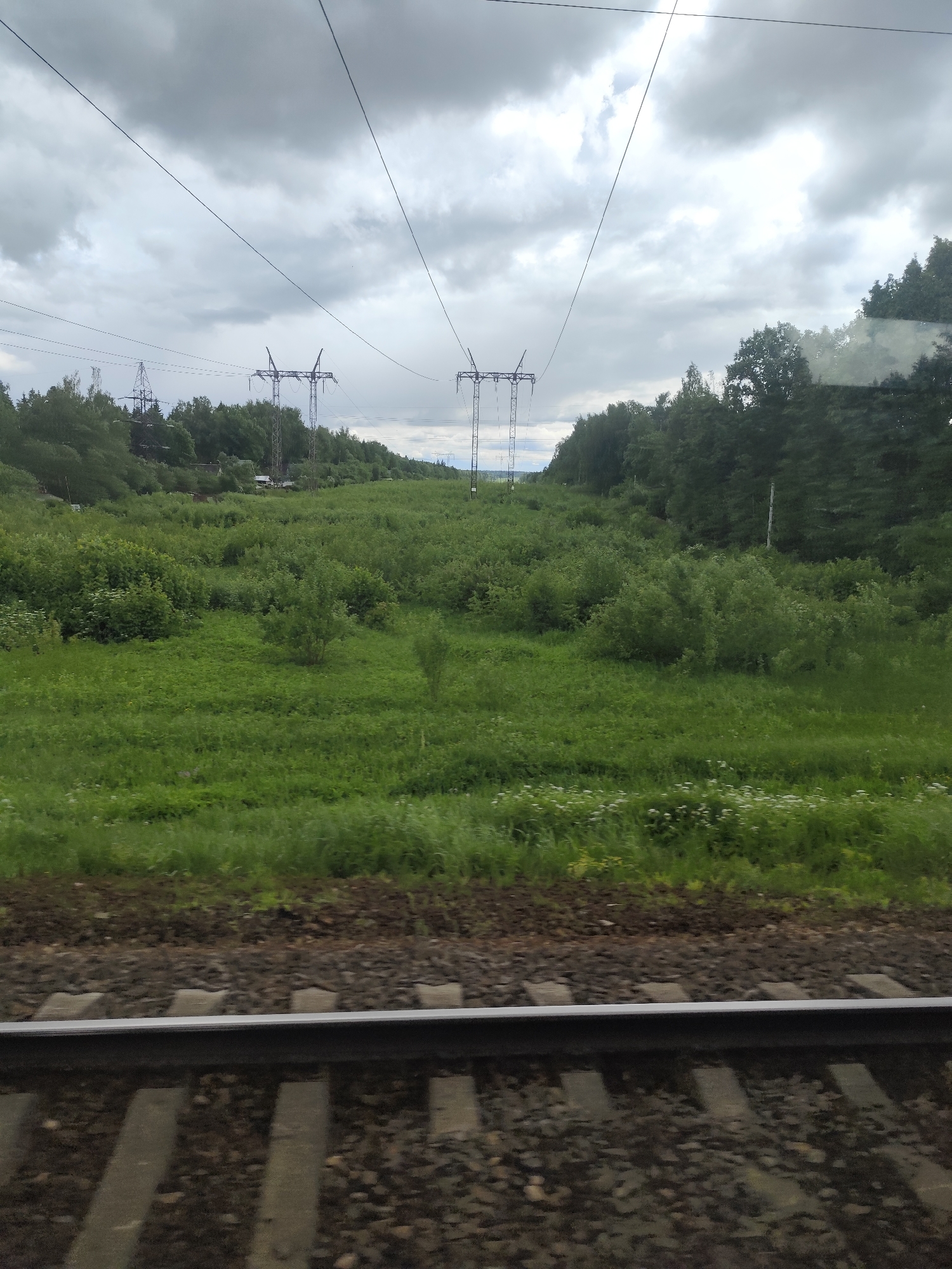 View from a train moving along a railway in Khotkovo, Russia. A green field stretches to a line of trees in the background, with three sets of electricity pylons in the distance. Overhead power lines run above the field. The sky is overcast with grey clouds. The railway tracks are in the foreground.