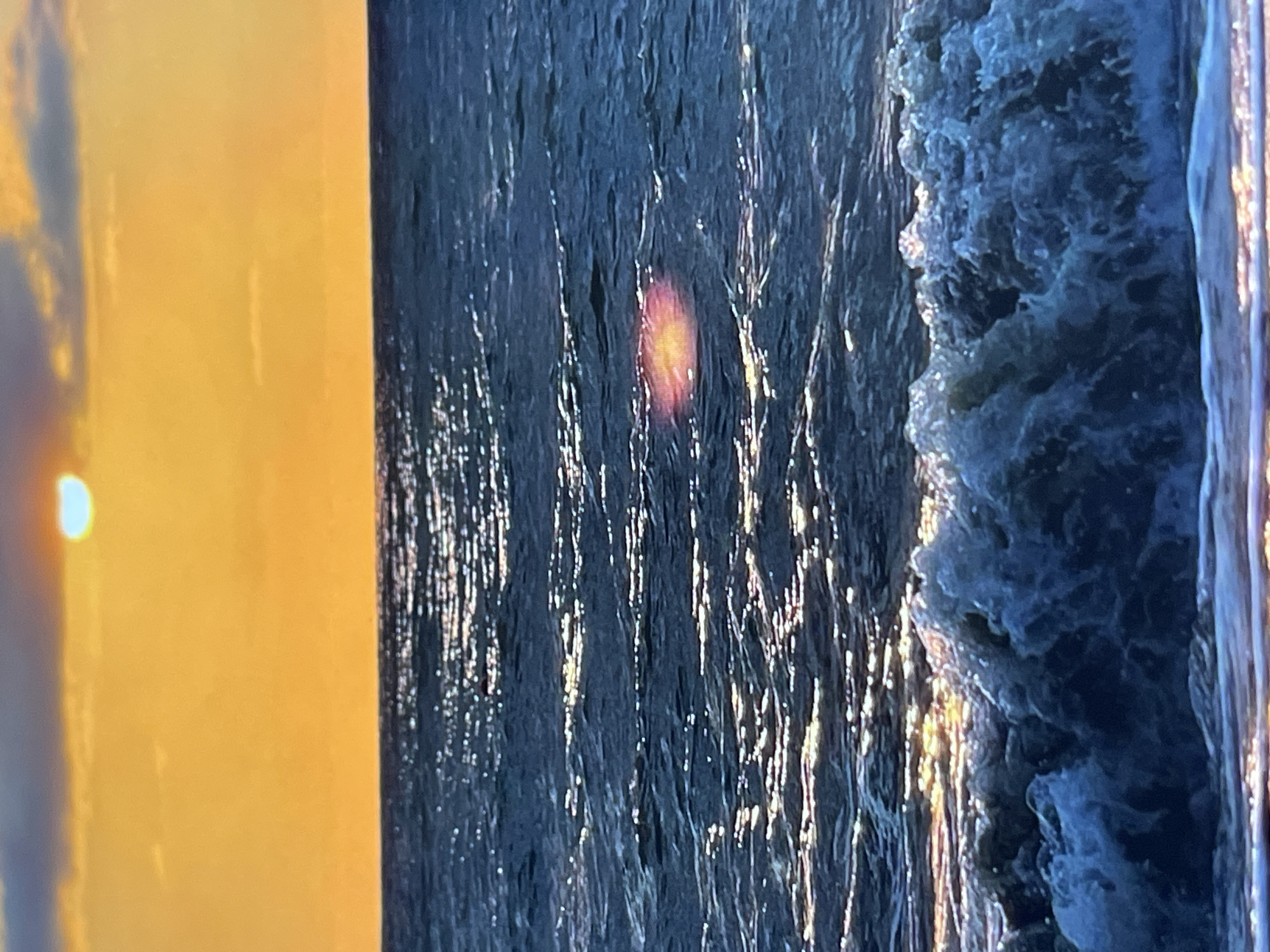 The image depicts a tranquil ocean scene at sunset. The sky is ablaze with warm orange and yellow hues, with a sliver of the sun visible near the top center, casting a bright glow. Wispy clouds are scattered across the sky, adding texture and depth. The ocean below is a deep blue, with gentle waves rolling towards the shore. Sunlight glints off the water's surface, creating shimmering reflections. In the foreground, a small wave crests, its white foam contrasting with the dark water. The overall impression is one of serene beauty and natural splendor. The location is identified as Unknown, Unknown.
