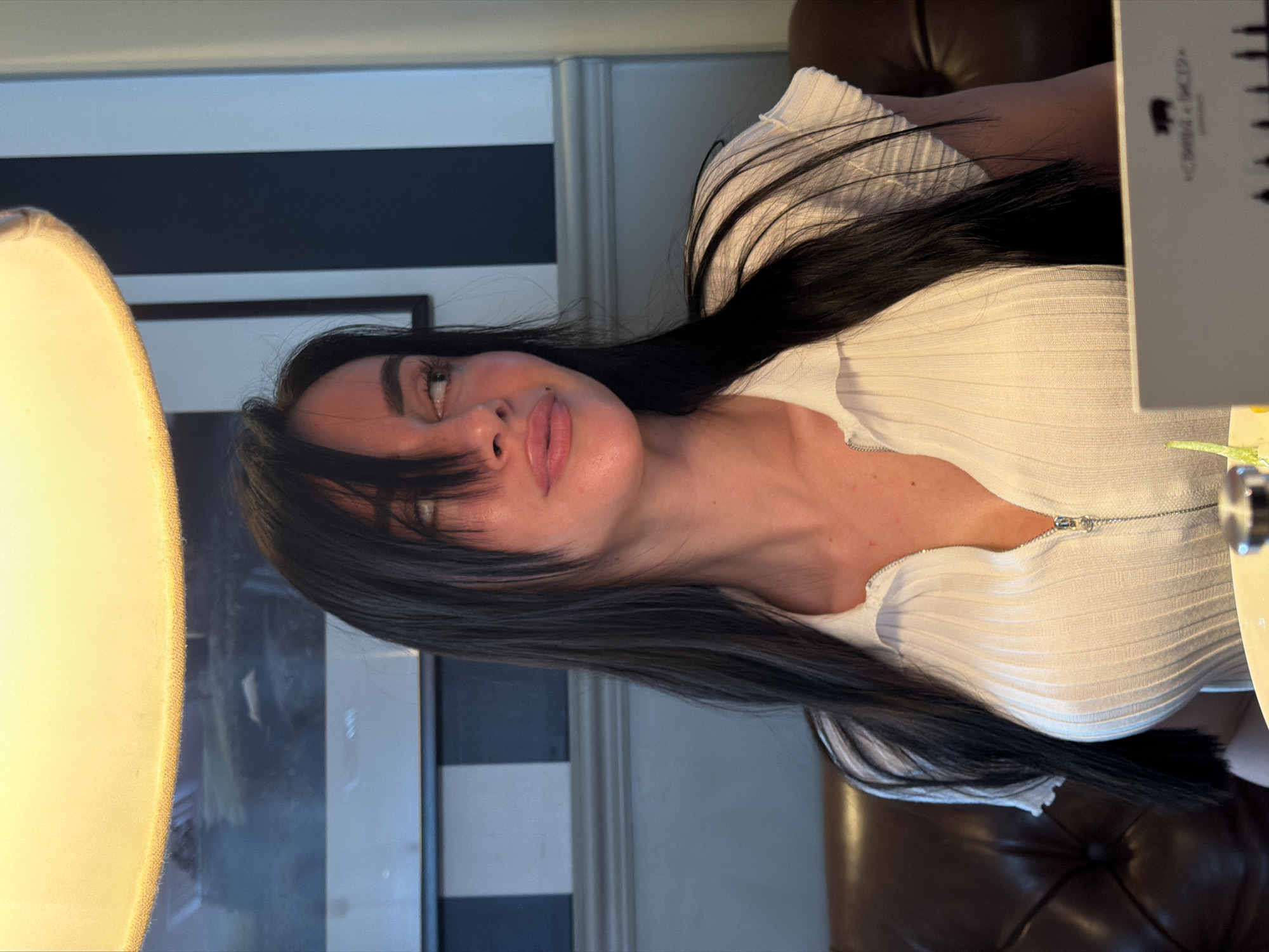 A woman with dark hair is wearing a white, ribbed shirt with a zipper closure. She is positioned indoors, seemingly at a restaurant or bar, given the presence of a menu in the foreground and a brown leather chair or booth in the background. The background includes a vertically striped wall and a framed picture. A bright light source is visible to the left. There is no verifiable information provided about a location in Krasnoyarsk, Russia.