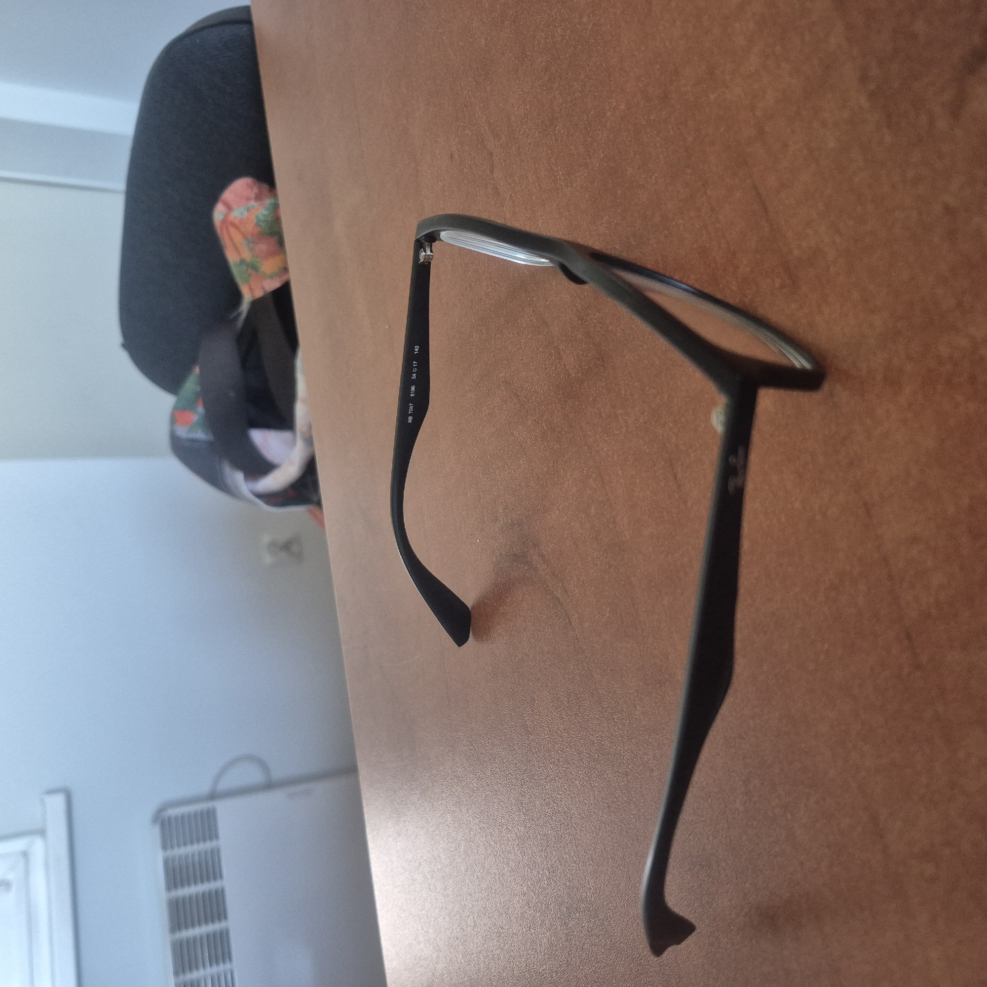 The image displays a pair of black eyeglasses lying on a brown, textured surface. The eyeglasses are positioned diagonally, with the lenses facing upwards and the arms extending downwards and to the right. The frame appears to be made of plastic. There is some text engraved on the inside of the right arm of the glasses.
On the left side of the image, a portion of a dark grey backpack is visible, with a colorful floral-patterned fabric peeking out from underneath. The background on the left is a pale blue wall, with a white object that resembles a radiator or vent.
The lighting suggests an indoor setting, possibly during the daytime given the brightness in the background. The brown surface the glasses are on could be a table or desk. There are no people visibly present in the frame, and the overall mood is neutral.
Visible text on the glasses: "RB 7047 5196 54 0 17 140".