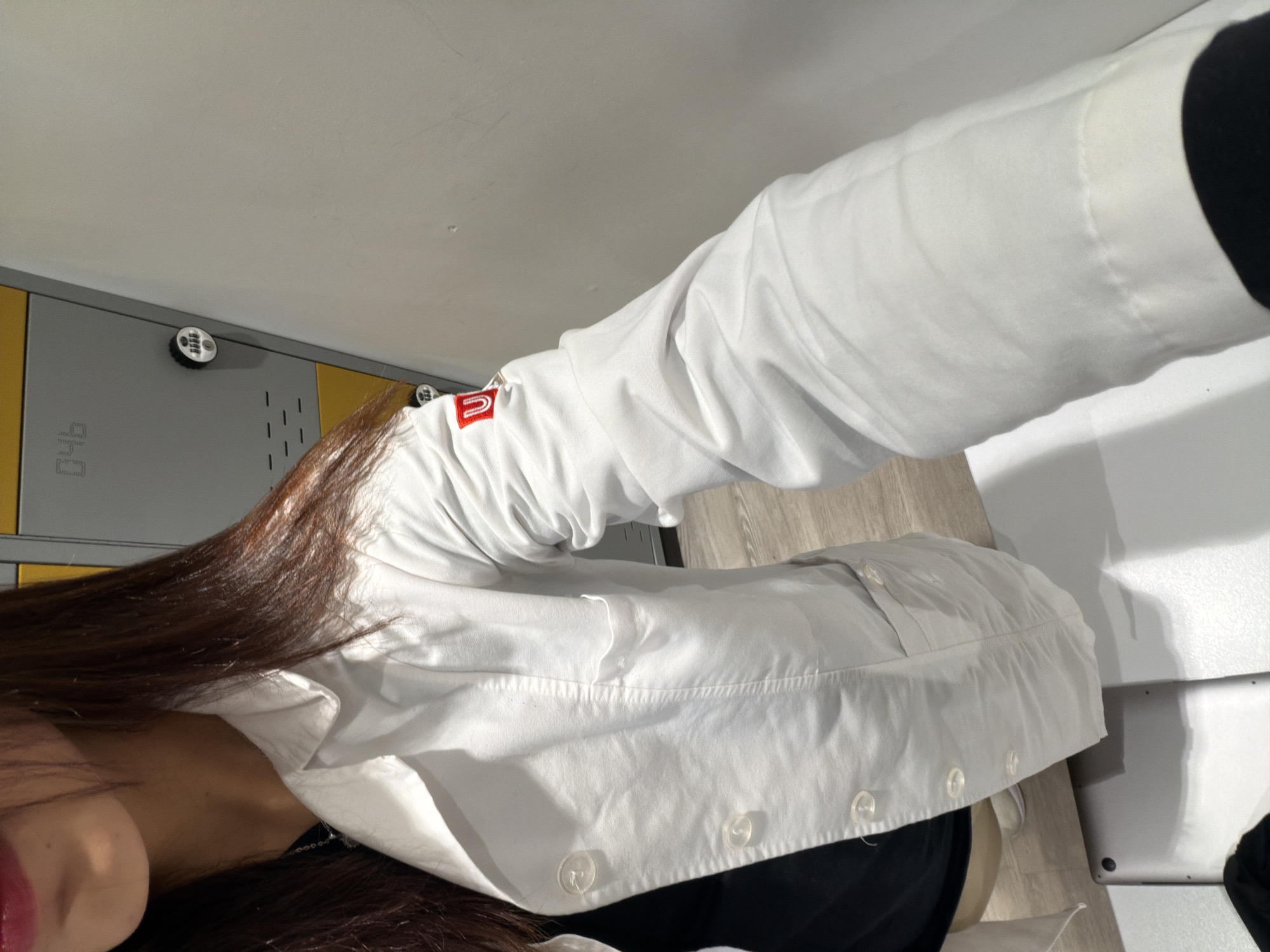 The image shows a close-up, waist-up view of a person wearing a white lab coat over a black top. The lab coat has clear buttons and two pockets, one on the chest and one lower down. On the left sleeve of the lab coat, there is a small red embroidered logo with a white "U" inside. The person has long, dark brown hair. In the background, there are lockers, some of which are yellow and grey, with the number "046" visible on one. The floor appears to be made of light-colored wood. A silver tablet or laptop is visible on a white surface in the lower left corner of the image. There is no visible indication of the location being in Bojacá, Colombia.
