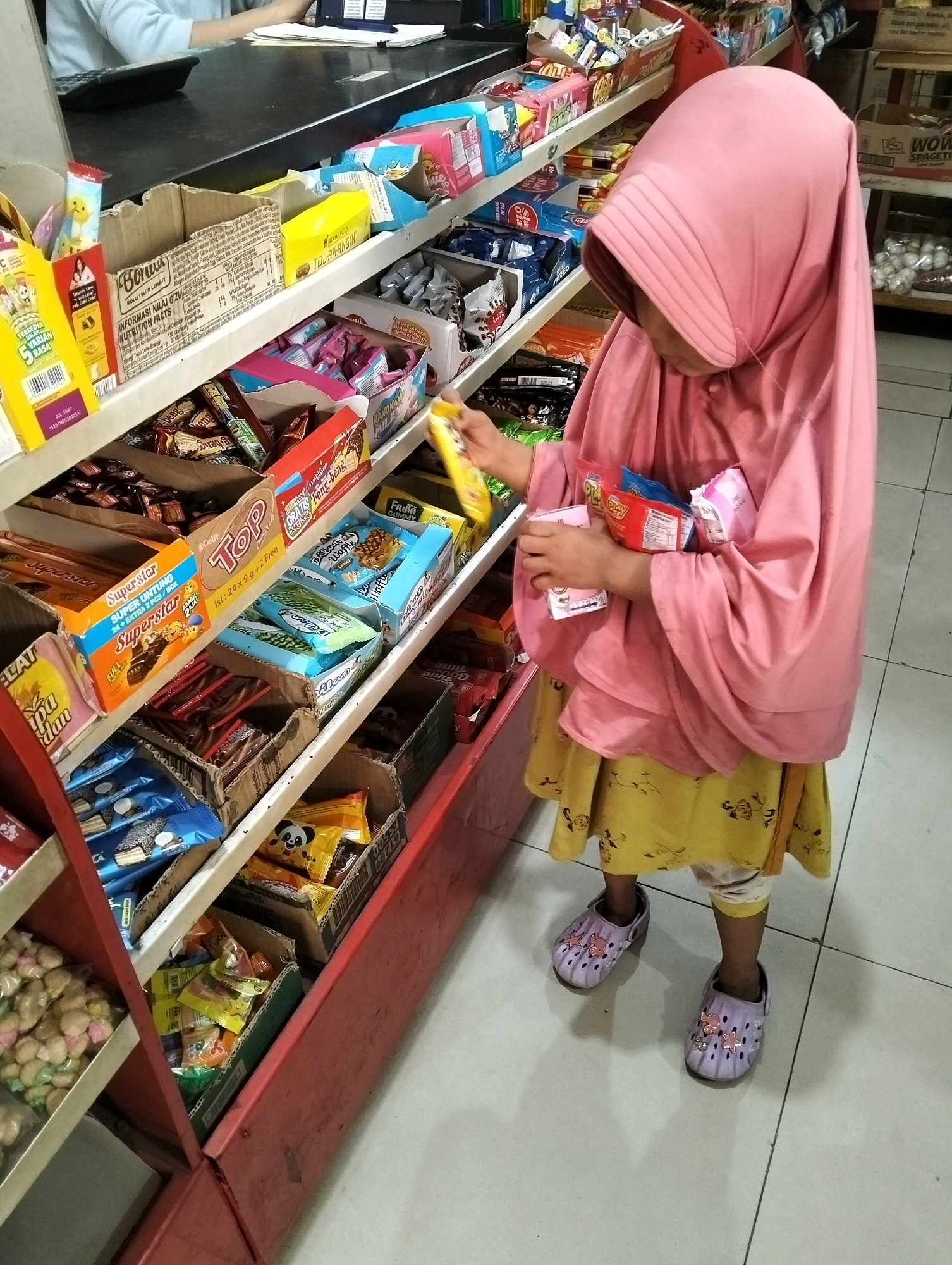 A young girl wearing a pink hijab and a yellow floral dress stands in an aisle of a convenience store in Cileungsi, Indonesia. She is holding several packaged snacks in her hands, reaching for another from a shelf. The shelves are filled with various branded snacks and candies. The floor is tiled and appears clean.