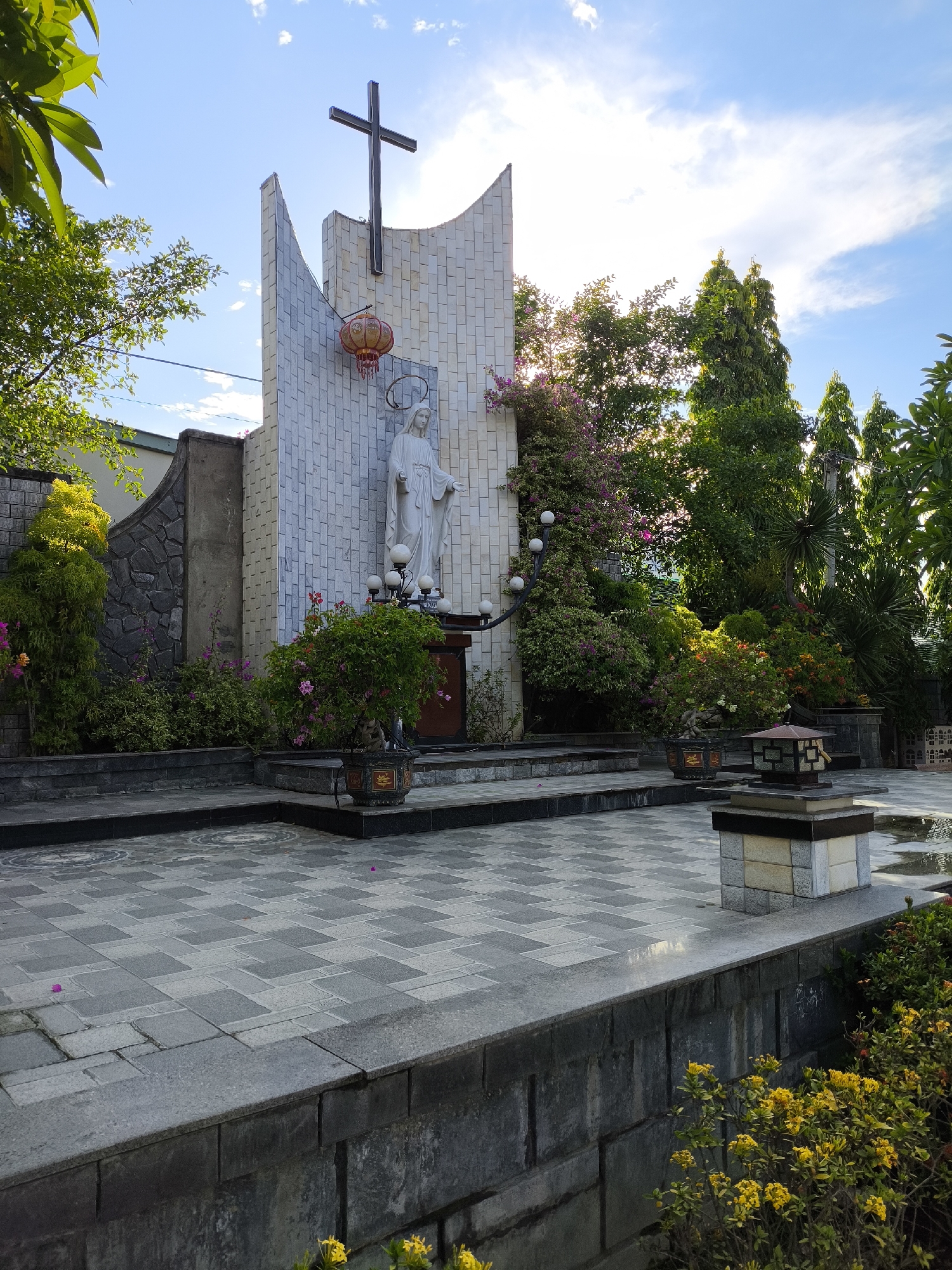 This media file captures a serene outdoor scene centered around a religious monument, likely a church or shrine, in Nha Trang, Vietnam. The primary subject is a white statue of a robed figure, possibly the Virgin Mary, standing on a raised platform. Above the statue, a dark, imposing cross is mounted on a textured, pale facade. The structure features a distinctive curved top and is adorned with a single orange lantern hanging to the left of the statue.
The setting is a courtyard paved with geometric grey tiles, some of which have circular etched patterns. The courtyard is bordered by raised stone platforms and surrounded by lush greenery. Various plants, including bushes with pink and orange flowers, and trees with vibrant green foliage, contribute to the tranquil atmosphere. A decorative stone lantern with a metal top stands on a low pedestal in the foreground.
The time of day appears to be late afternoon or early evening, with the sun casting a warm glow and creating some lens flare visible in the upper right portion of the image. The sky is a clear blue with scattered white clouds, suggesting pleasant weather. The overall mood is peaceful and contemplative, amplified by the natural elements and the solemn religious imagery. There is no visible text in the image. The combination of the religious monument, the manicured gardens, and the architectural style suggests a place of worship or a memorial in a tropical or subtropical location.