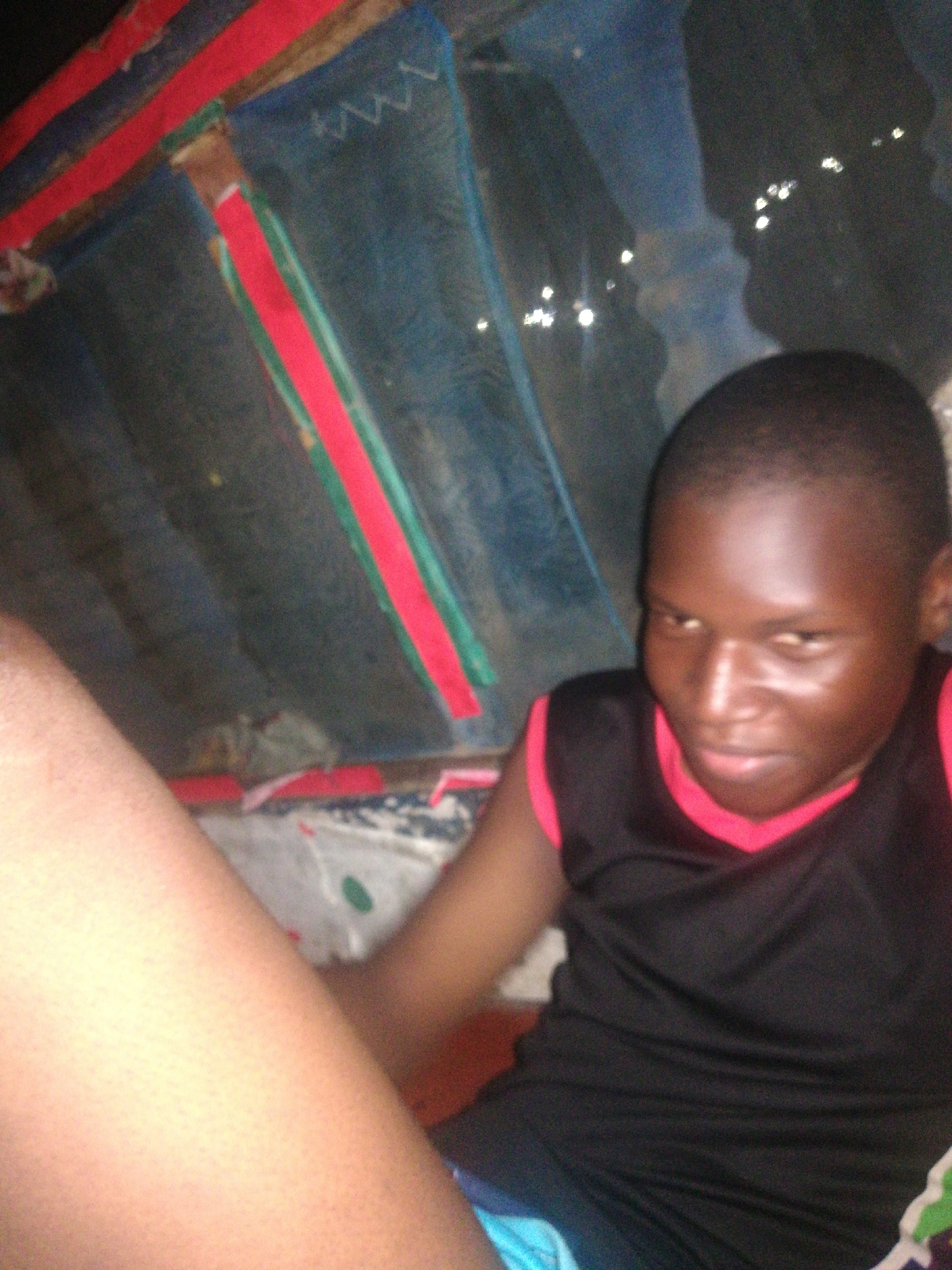 The image appears to be taken inside a room in Lagos, Nigeria. A dark-skinned person, possibly a young man, is visible. He is wearing a black sleeveless shirt with pink trim. Part of a leg is visible in the foreground. The background features blue and red striped fabric. A sheet with a pattern of white, blue, and green is visible underneath. Additional bright, out-of-focus spots can be seen in the top right, possibly indicating distant lights.