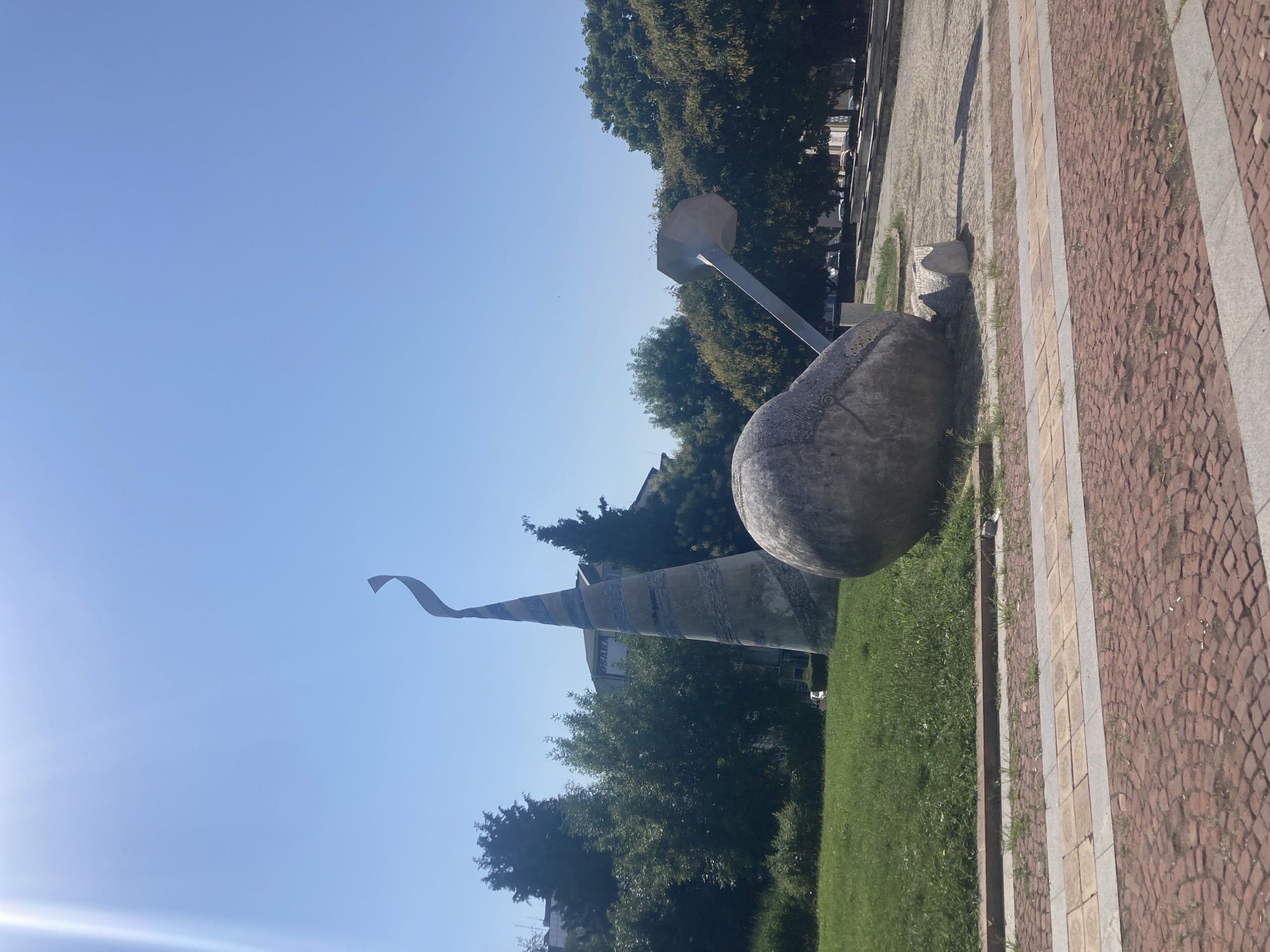 The image depicts a sunny outdoor scene in Burgas, Bulgaria, featuring a contemporary sculpture in a public space. The sculpture consists of two main elements: a large, rounded, stone-like object with a metallic shaft extending from it, resembling a stylized hammer or pestle, and a tall, conical, segmented structure that tapers to a point and curves at the top. The conical element appears to be made of concrete or a similar material, decorated with horizontal bands of varying colors and textures. The ground in the foreground is paved with red brickwork laid in a pattern, bordered by a strip of lighter colored paving stones and a concrete curb. Beyond the paved area, there is a patch of green grass with some sparse weeds. In the background, a line of mature trees with full green foliage forms a backdrop, interspersed with glimpses of buildings. The sky is a clear, bright blue with a sun flare visible in the upper left corner. There are no people or animals visible in the frame. The overall impression is one of a public park or plaza designed for artistic display.