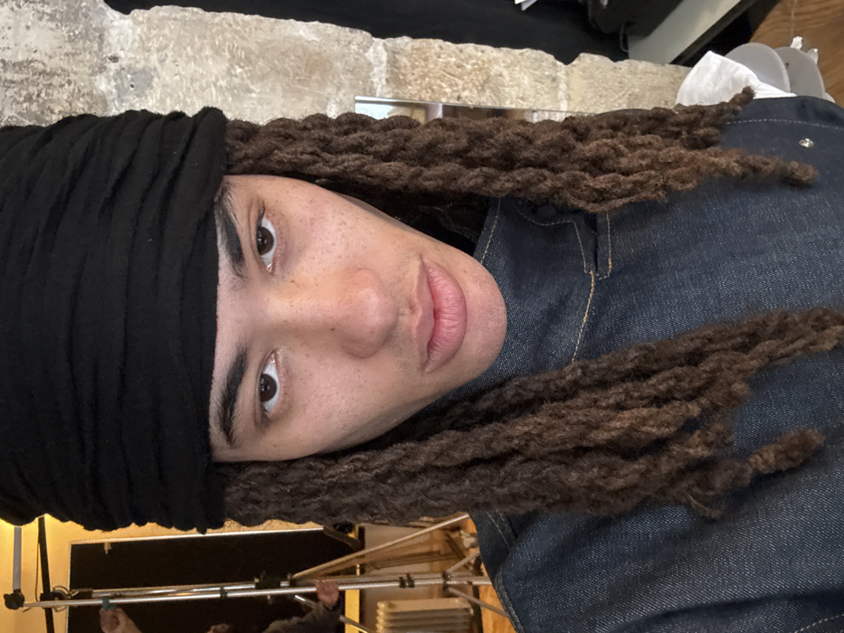 A portrait-style image focuses on a person with medium-dark skin, wearing a black fabric headwrap and long, dark brown dreadlocks or braids. The individual looks directly at the camera with dark eyes and has visible freckles across the face. A dark blue denim-like jacket or shirt with light-colored stitching is worn.
The indoor environment suggests a photoshoot setting. To the left of the subject, studio equipment is visible, including a metal light stand with a boom arm, a portion of a white reflector, and a human hand. Below this, a white radiator is present against a light-colored wall. To the right, a textured, light-colored wall, possibly stone or brick, dominates the background, with a dark vertical object and a stack of white circular items alongside a crumpled white cloth visible at the lower right edge.
Based on the visual evidence, the specific location within the city of Paris, France, cannot be determined. The scene appears to be an interior space, likely a studio or a room adapted for photography.