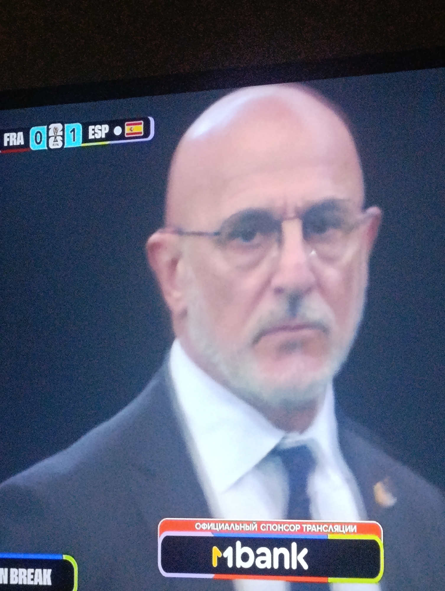 The image is a medium shot of a man with a bald head and glasses. He is wearing a suit and tie. In the background, a sports score is displayed on a screen showing "FRA 0 1 ESP" and the Spanish flag. Below the man's chest is a rectangular banner with text that appears to be in Russian and a logo. The environment is dark and appears to be indoors, possibly in a stadium or a television studio. The location in Osh City, Kyrgyzstan, is not discernible from the image.
