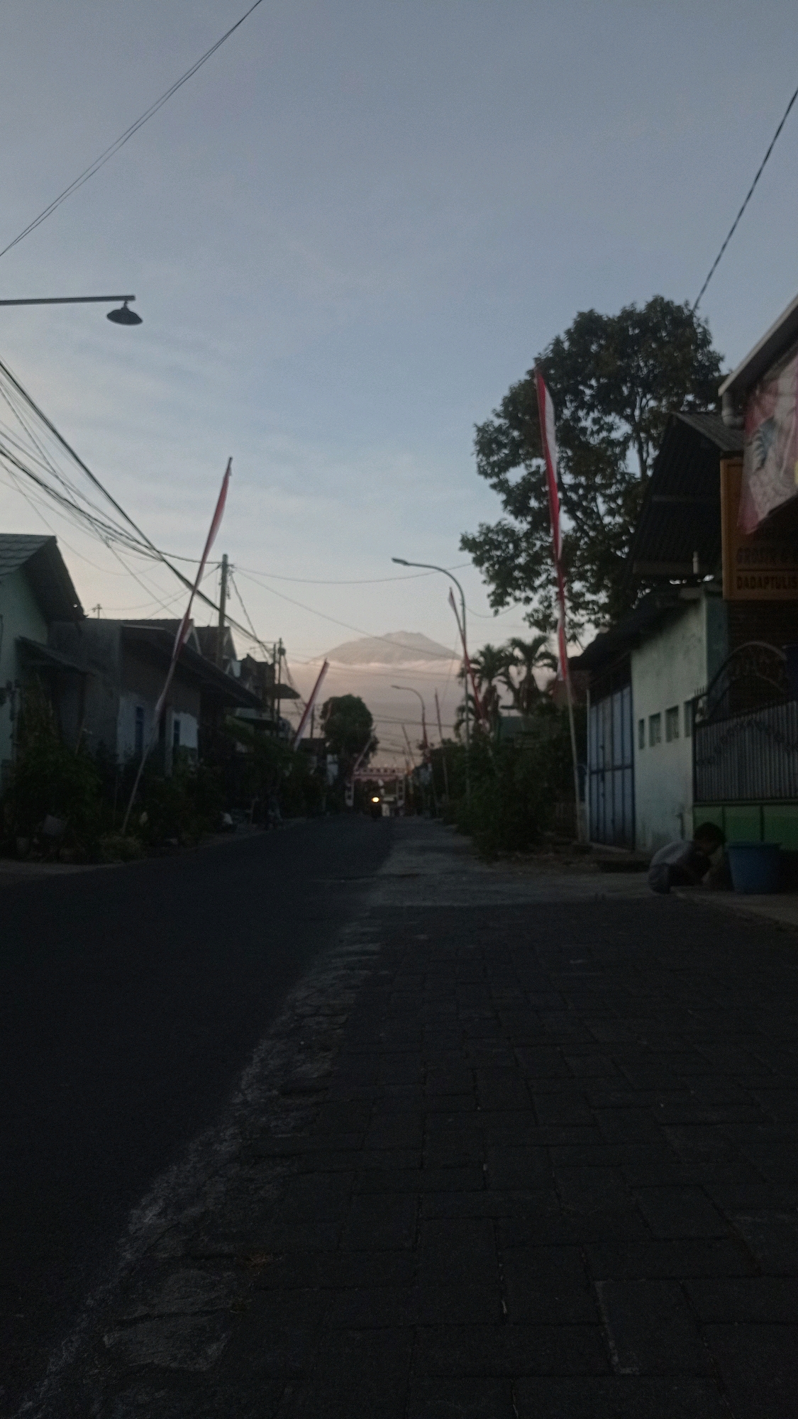 The image depicts a quiet, paved street in Batu, Indonesia, likely captured at dawn or dusk due to the soft, diffused light. The street is flanked by modest houses and trees, with numerous electrical wires crisscrossing overhead. Two Indonesian flags, red and white, are prominently displayed on poles along the roadside, suggesting a national holiday or a patriotic display. In the distance, a large mountain shrouded in mist is visible, hinting at the natural beauty surrounding the city. A person is seen crouching on the right side of the street, near a blue bin. The overall atmosphere is serene and tranquil, with a sense of everyday life in a small Indonesian town.