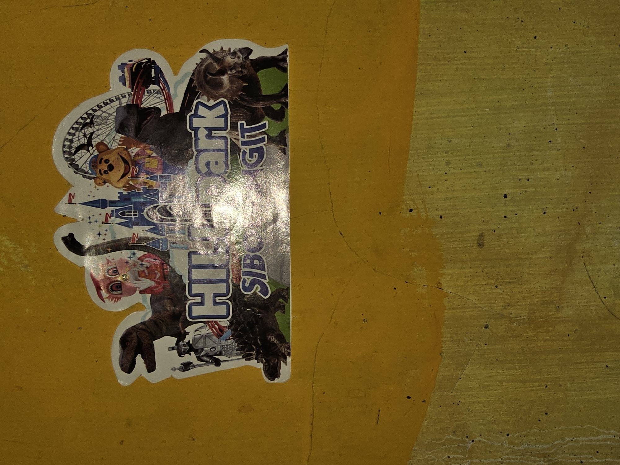 The image shows a sticker on a yellow concrete wall. The sticker is a colorful cartoon depiction of a theme park. It features various characters and attractions, including dinosaurs, a teddy bear mascot, a castle, a Ferris wheel, and a roller coaster. The text on the sticker reads "HI Park SIBU GTT". The wall itself appears weathered and cracked, with some dirt and debris visible. The location in the city of Payagaboh, Indonesia is not evident from the image.