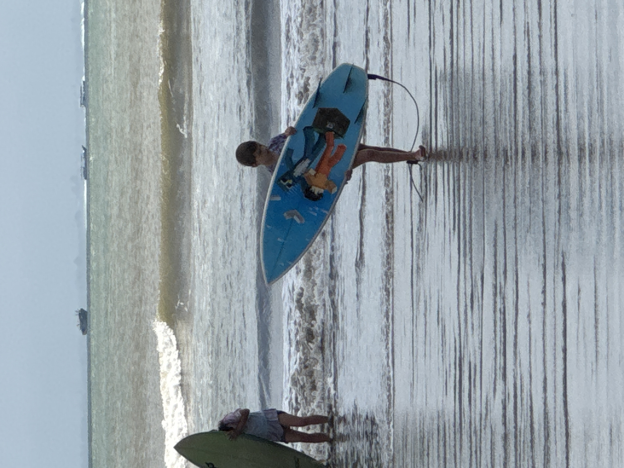 In Kuta Selatan, Indonesia, a child with short brown hair stands on a wet sandy beach, holding a blue surfboard featuring cartoon graphics of people and what appears to be a suitcase. The child is wearing shorts and a t-shirt. To the left, another person with dark hair is partially visible, holding a green surfboard and standing in shallow water. The ocean has waves, and in the distance, there are several boats, including a larger cargo ship. The sky is overcast.