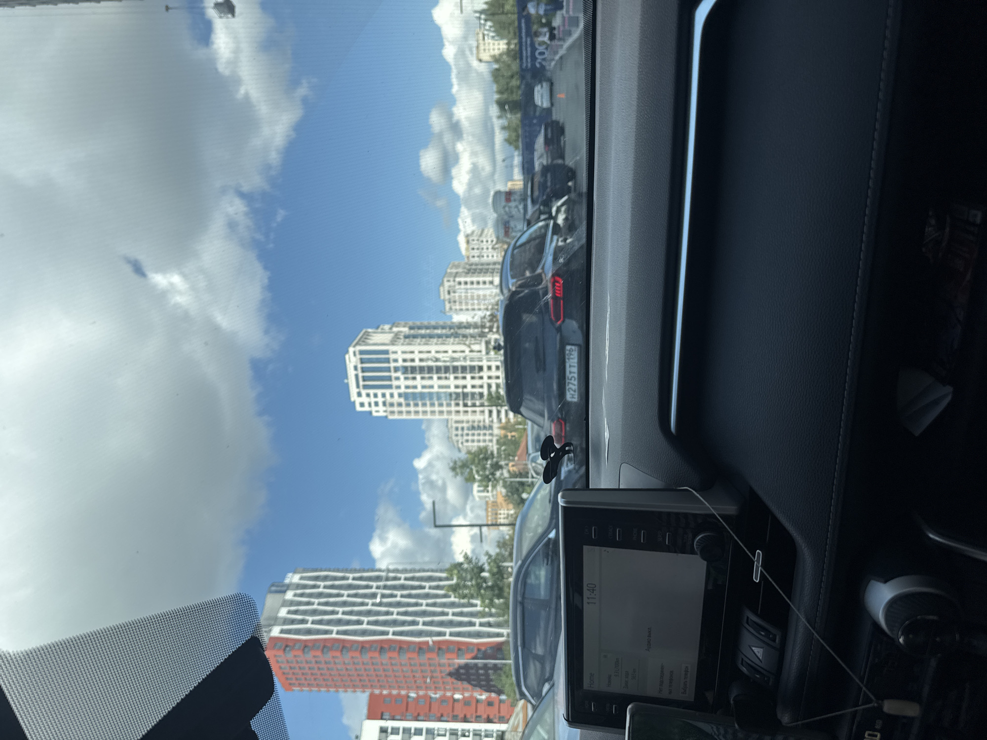 The image captures a view from inside a car, looking forward through the windshield at a traffic jam. The dashboard of the car is visible in the foreground, with a navigation screen displaying the time "11:40". Beyond the car, a city street is lined with modern buildings, including a tall red and white patterned skyscraper on the left and a white skyscraper with numerous windows on the right. Several cars are stopped in traffic, including a dark-colored SUV with a license plate that reads "P 275 TT 196". The sky is blue with fluffy white clouds. The overall scene suggests a typical urban environment during the daytime.