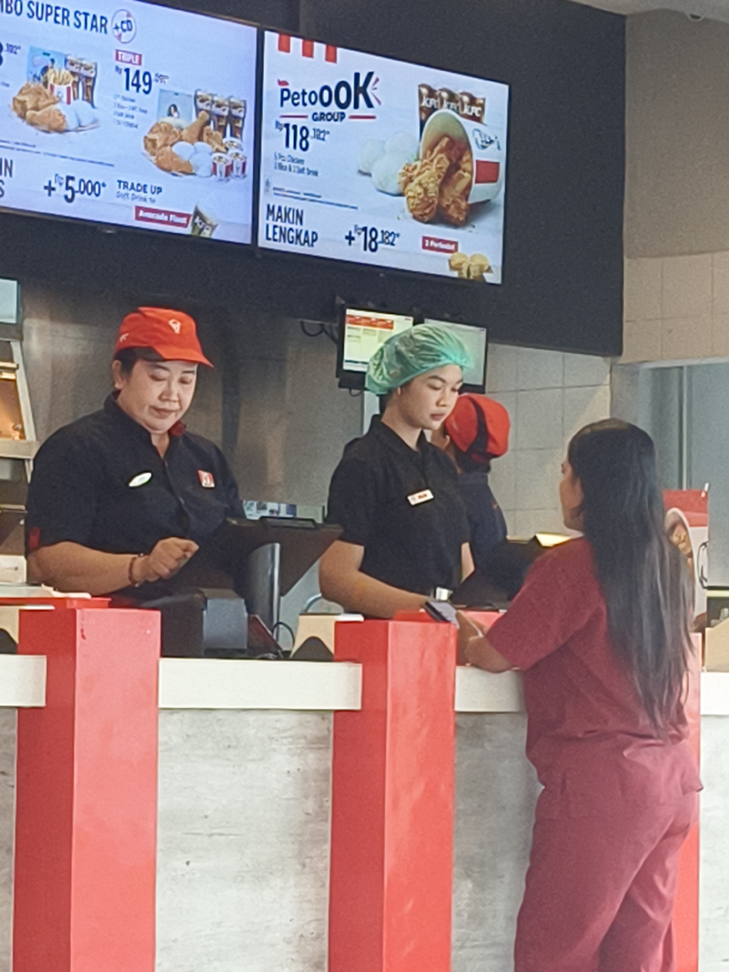 The image depicts the interior of a KFC restaurant in Denpasar, Indonesia. Two female employees are visible behind a counter. The employee on the left, wearing a red KFC-branded cap and a dark uniform, is operating a digital tablet. The employee on the right, wearing a green hairnet and a dark uniform, is facing a customer whose back is to the camera, wearing a red uniform. Above the counter, digital menu boards display various KFC promotions. The environment appears to be a typical fast-food restaurant setting with red and white counter accents.