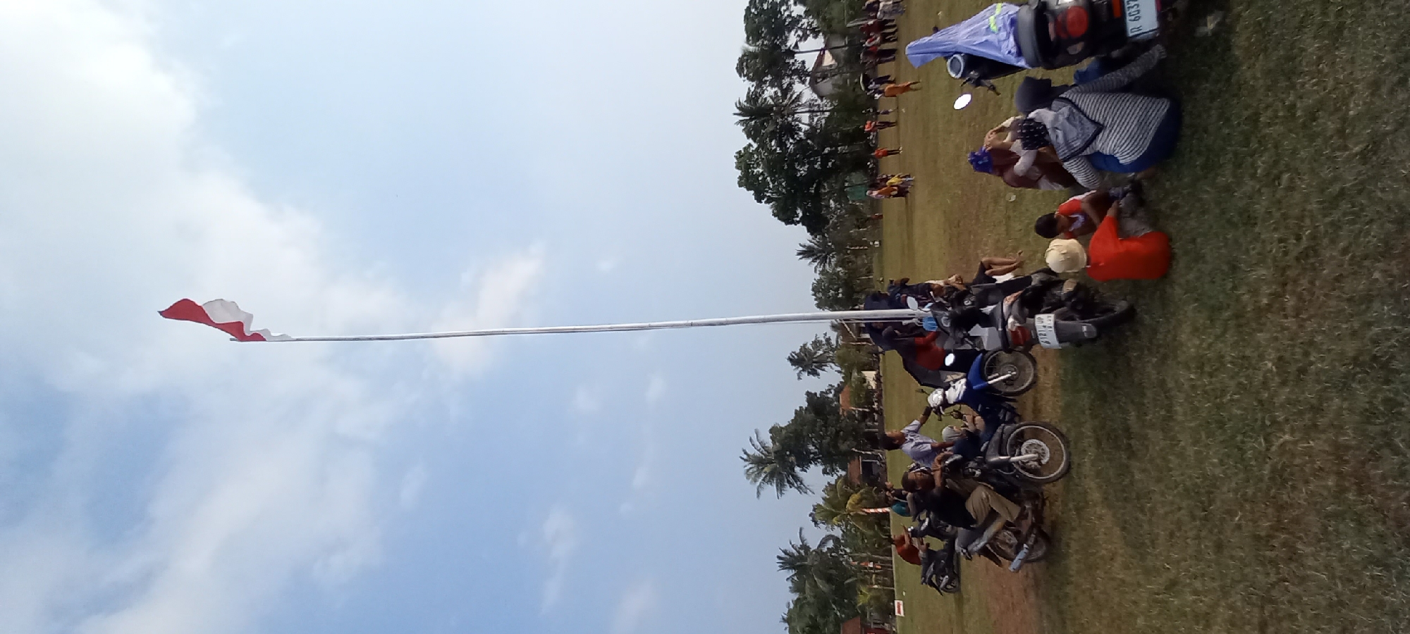 In Sidamulya, Indonesia, a group of people are gathered on a grassy field, with a tall flagpole standing prominently in the center. The Indonesian flag, red and white, is visible at the top of the flagpole. Several motorcycles are parked around the gathering, suggesting a local event or assembly. The people are dressed casually and appear to be observing or participating in an activity. The environment is outdoors, with trees and buildings visible in the background under a clear sky with some clouds. The scene suggests a community gathering or a public ceremony taking place in a rural or semi-rural area.