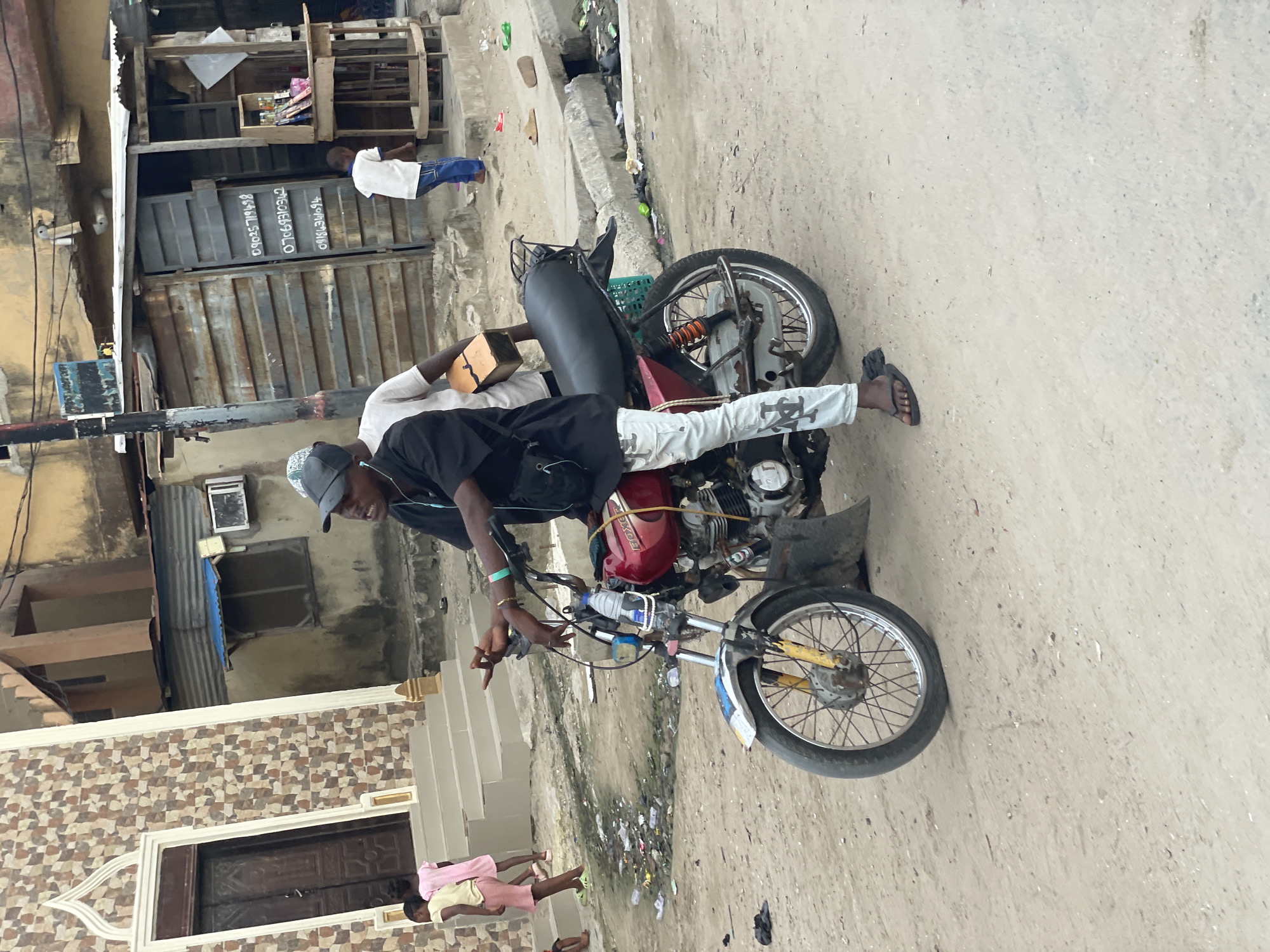 The image, rotated 90 degrees clockwise, captures an outdoor street scene in Ado, Nigeria, during daylight hours. In the foreground, two men are positioned around a red "BOXER" brand motorcycle. One man, wearing a black cap, black shirt, and light-colored trousers with an "NY" logo, is seated on the motorcycle, holding the handlebars. Behind him, another man in a white shirt and dark trousers stands, holding a brown rectangular box.
The setting appears to be a bustling, somewhat weathered urban or semi-urban area. To the left, a building features a decorative tiled facade with an ornate wooden door, in front of which two children, one in pink and one in yellow attire, are standing. Further left and in the background, there's another building with corrugated metal siding and an exposed window, beneath which a small, makeshift wooden stall or shop displays various items. A child in a white shirt and blue trousers stands near this stall. The ground is an unpaved, dusty path with scattered debris, including plastic bottles and other trash.
Visible text on a corrugated metal gate includes phone numbers and names: "TO OLUWAGBEMIGA 07069145964" and "08064860640". The overall atmosphere suggests a casual, everyday moment in a local neighborhood.