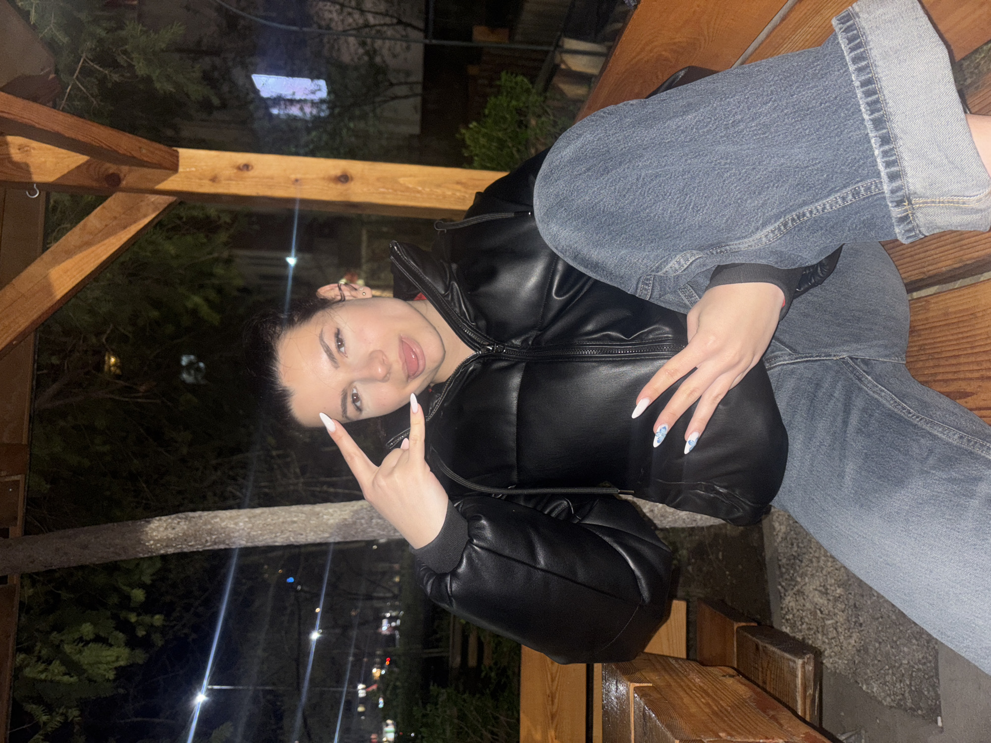 A young woman is sitting on a wooden bench outdoors at night. She is wearing a black puffer jacket and blue jeans with cuffed hems. She is making a hand gesture, sticking out her tongue, and posing for the photo. There are trees and city lights visible in the background. Based on the background context, this location seems to be in the city of Plovdiv, Bulgaria.