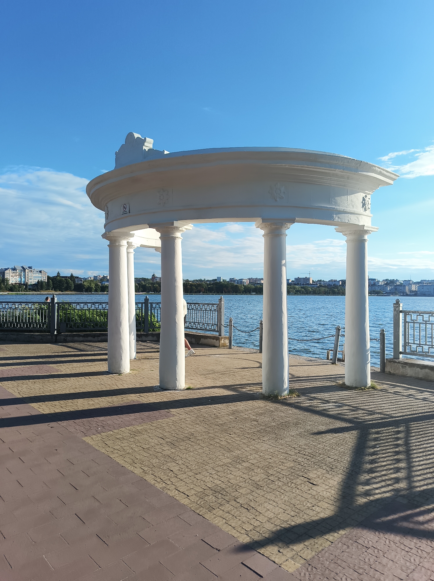 The image depicts a picturesque waterfront scene, likely the Ternopil Pond in Ternopil, Ukraine. In the foreground, a paved walkway transitions from a darker reddish-brown material to a lighter tan brick pattern. Dominating the mid-ground is a white, open-air rotunda supported by four substantial white columns. The rotunda features a decorative, curved roofline with intricate detailing. Beyond the rotunda, a body of blue water stretches across the frame, reflecting the clear blue sky dotted with a few wispy clouds. A metal fence lines the edge of the walkway separating it from the water. In the background, the cityscape of Ternopil is visible, characterized by buildings of various sizes and architectural styles, interspersed with lush green trees. The overall impression is one of a tranquil, scenic public space bathed in natural light.