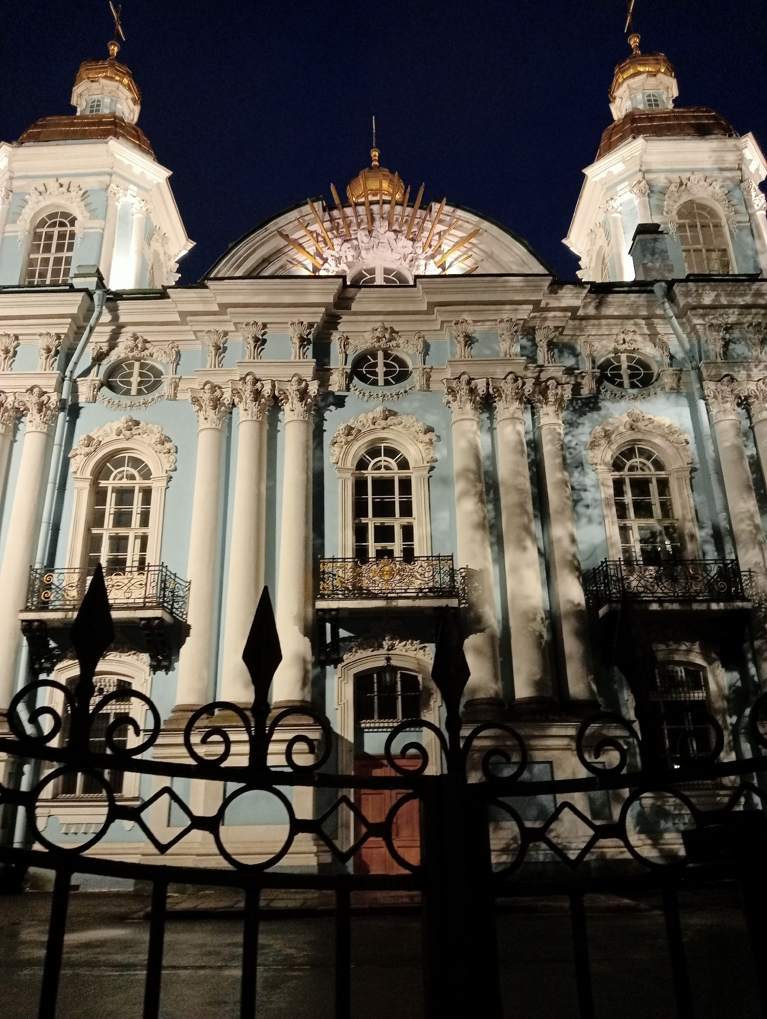 This is a nighttime, low-angle, vertical shot of the St. Nicholas Naval Cathedral in Saint Petersburg, Russia. The ornate Baroque facade of the light blue cathedral is illuminated, highlighting its white decorative elements, columns, and arched windows. Two gold-domed towers rise on either side of the central structure, each topped with a cross. A decorative golden sunburst design is visible at the peak of the main facade. In the foreground, a black ornate wrought-iron fence with scrollwork and diamond shapes partially obscures the view of the cathedral's lower level, which includes windows and a reddish-brown door. The sky is a deep, dark blue.