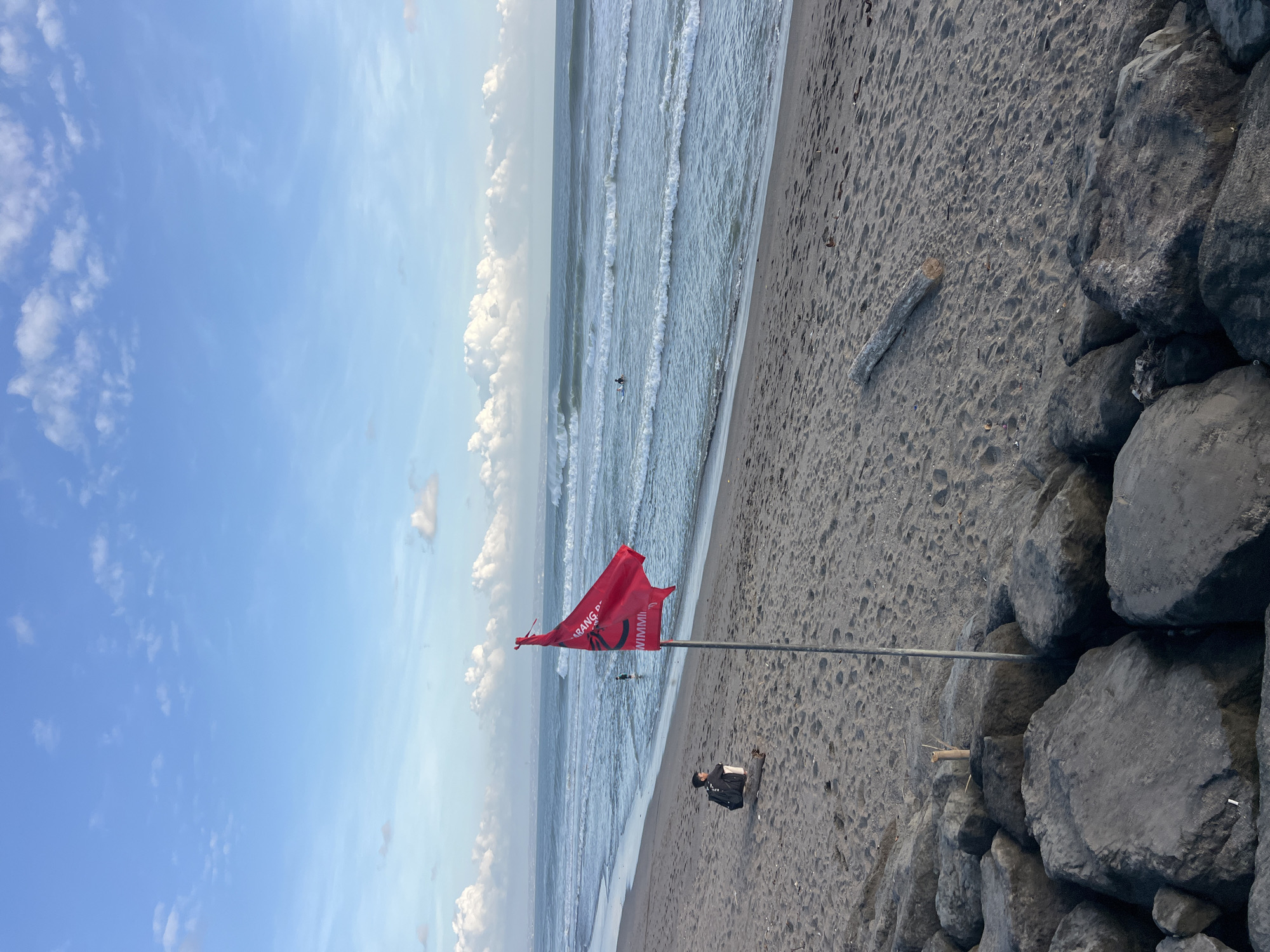 A red flag with white text that reads "DILARANG BERENANG" (which translates to "NO SWIMMING" in Indonesian) is planted in the sand on a beach in Mengwi, Indonesia. The flag is partially obscured by its pole and is flapping in the wind. In the background, the ocean has moderate waves, and a few surfers are visible in the water. A person is sitting on a log on the sandy beach, facing the ocean, with a backpack next to them. The beach is lined with large, dark rocks in the foreground. The sky is blue with scattered white clouds.