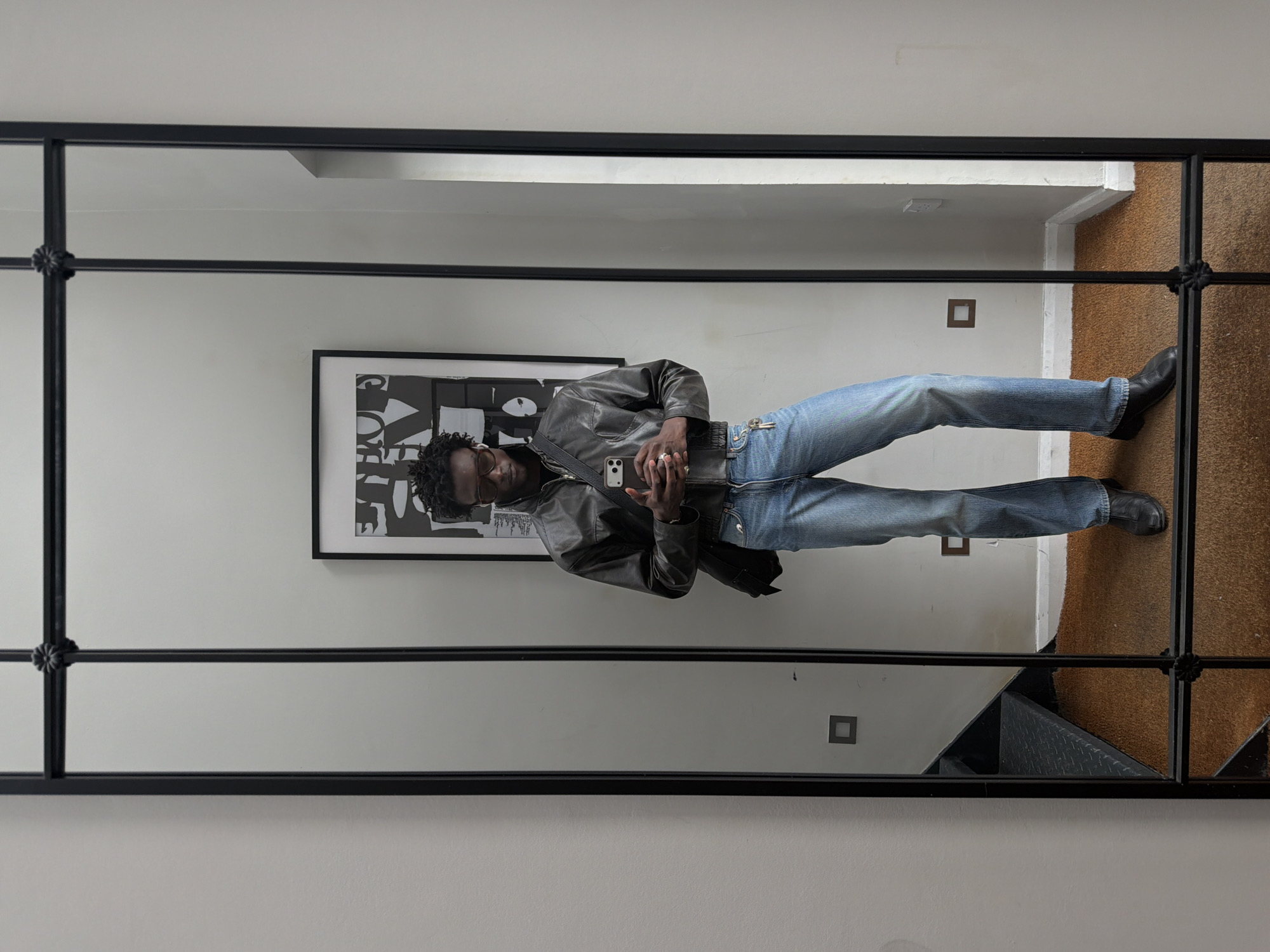 A reflection in a black-framed mirror shows a black male indoors, likely in London, UK, based on architectural style. The man wears a black leather jacket, light blue jeans, and black shoes. He's holding a mobile phone. Behind him, the wall features a black-and-white framed artwork. The floor is carpeted in brown. There are light switches on the wall.