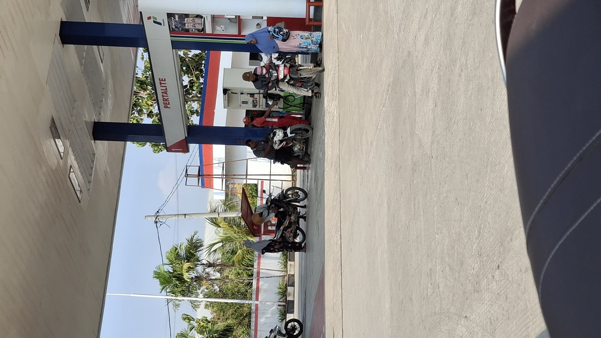This image depicts a scene at a Pertamina gas station in Pucakwangi, Indonesia. Several individuals are present, interacting with motorcycles and the gas pumps. On the left, a person in dark clothing with a headscarf stands next to a parked motorcycle. Further right, two men are attending to motorcycles near the pumps. One man in a red shirt is operating a gas pump. Another man in a gray shirt is bent over a white motorcycle. In the center, a person wearing a yellow head covering is seated on a motorcycle. To the right, a man in a gray shirt and cap is on a motorcycle, and a woman in a blue hijab and patterned dress is standing nearby, holding a blue bag. The gas station features a white and blue awning supported by blue pillars, with red and white accents on the building. Palm trees and other greenery are visible in the background, along with utility poles and wires. The ground is paved concrete, and the sky is clear and blue. The overall impression is a functional, outdoor retail environment.