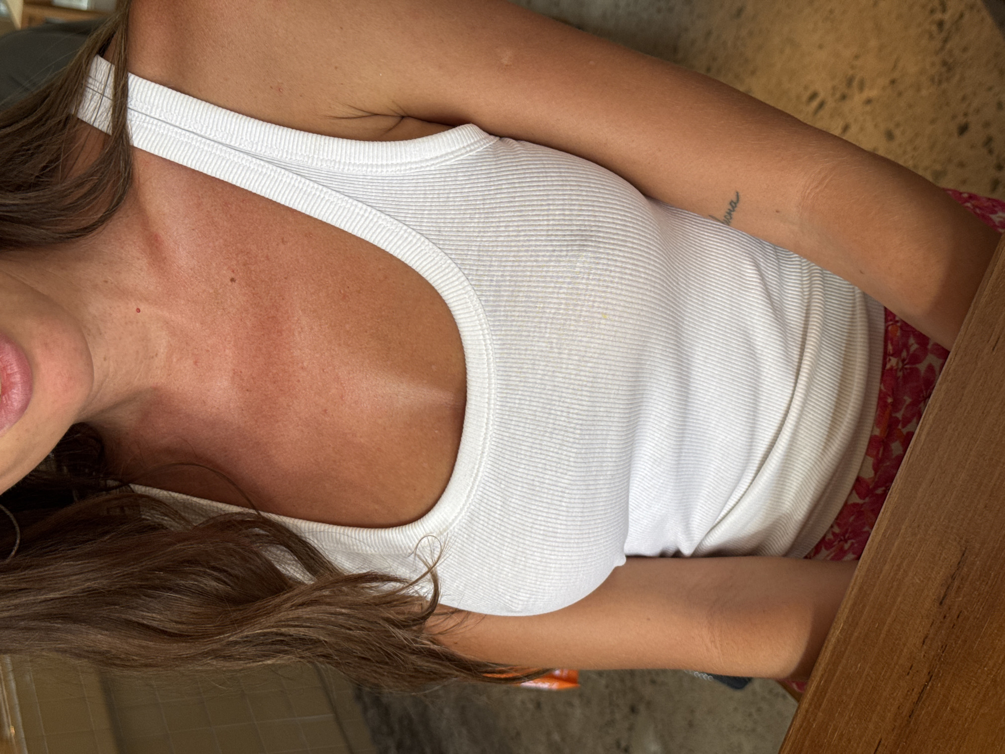 The image is a close-up, slightly angled overhead shot of a woman wearing a white ribbed tank top. Her right arm, with a tattoo on the forearm, is visible. The left side of her face, including part of her mouth, is also visible, along with strands of long, brown hair. She is seated at a wooden table, and a floral-patterned garment is visible beneath the tank top. The background is blurred and suggests an indoor setting. The location of North Kuta, Indonesia, is not discernible from the visual information provided in the image.