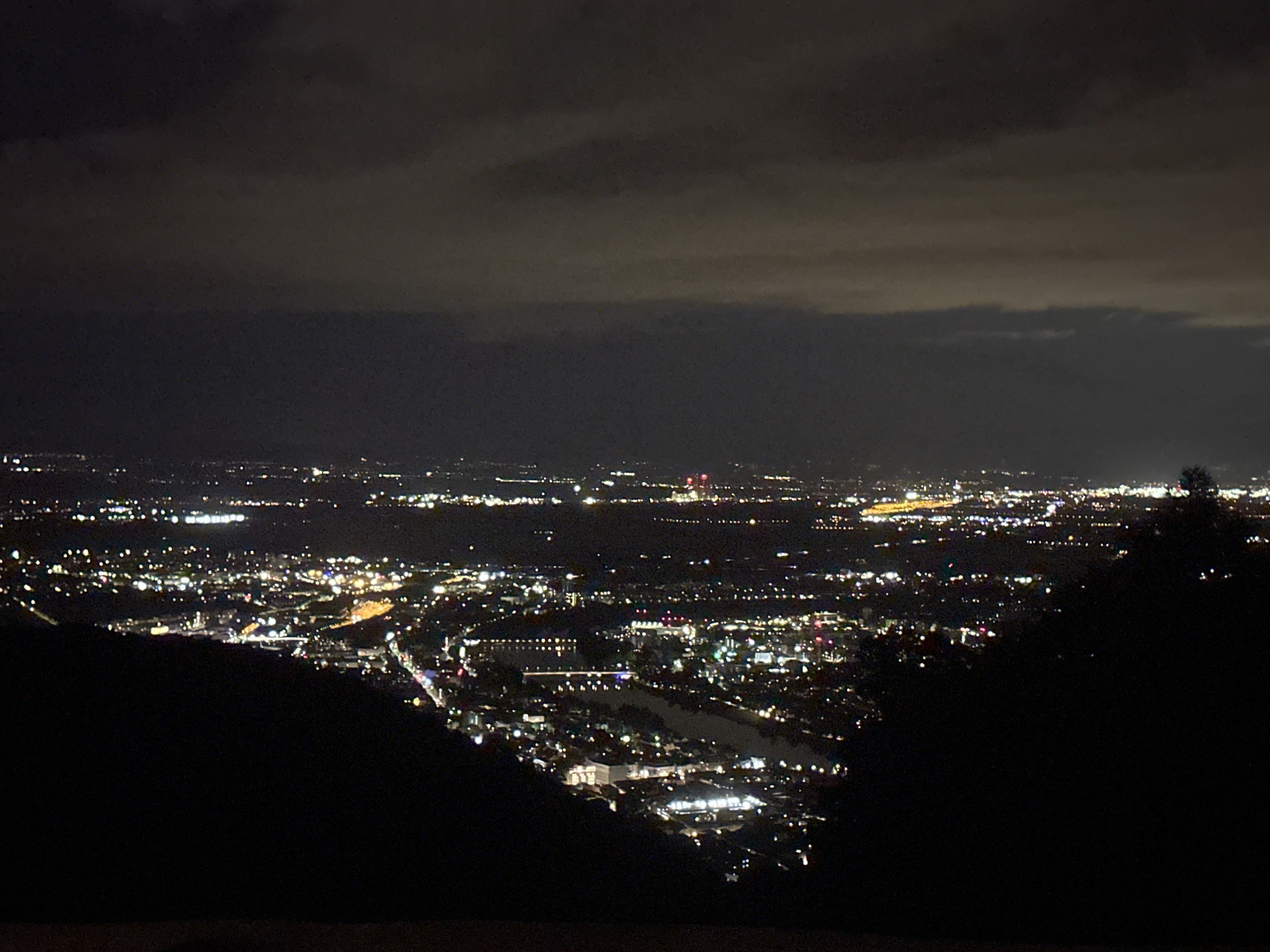 This is a nocturnal photograph presenting an expansive view of the city of Heidelberg, Germany, observed from an elevated vantage point. The upper half of the frame is dominated by a dark, heavily overcast sky, featuring distinct layers of grey clouds that subtly reflect the ambient light from the city below. The central and lower portions of the image display a wide urban spread, characterized by a dense array of artificial lights. These lights, primarily white and yellow with occasional red and blue sources, illuminate the city's infrastructure, defining roads, buildings, and residential areas. A prominent winding body of water, identifiable as the Neckar River, traverses the cityscape, reflecting the urban luminosity on its surface. In the distant background, brighter clusters of lights and a notable pair of red structures are visible. The immediate foreground, particularly in the lower-center and right, is occupied by a dark, silhouetted landmass, indicative of a hillside or mountain, which frames the illuminated city. No discernible individuals, animals, or specific actions are present in the image.
