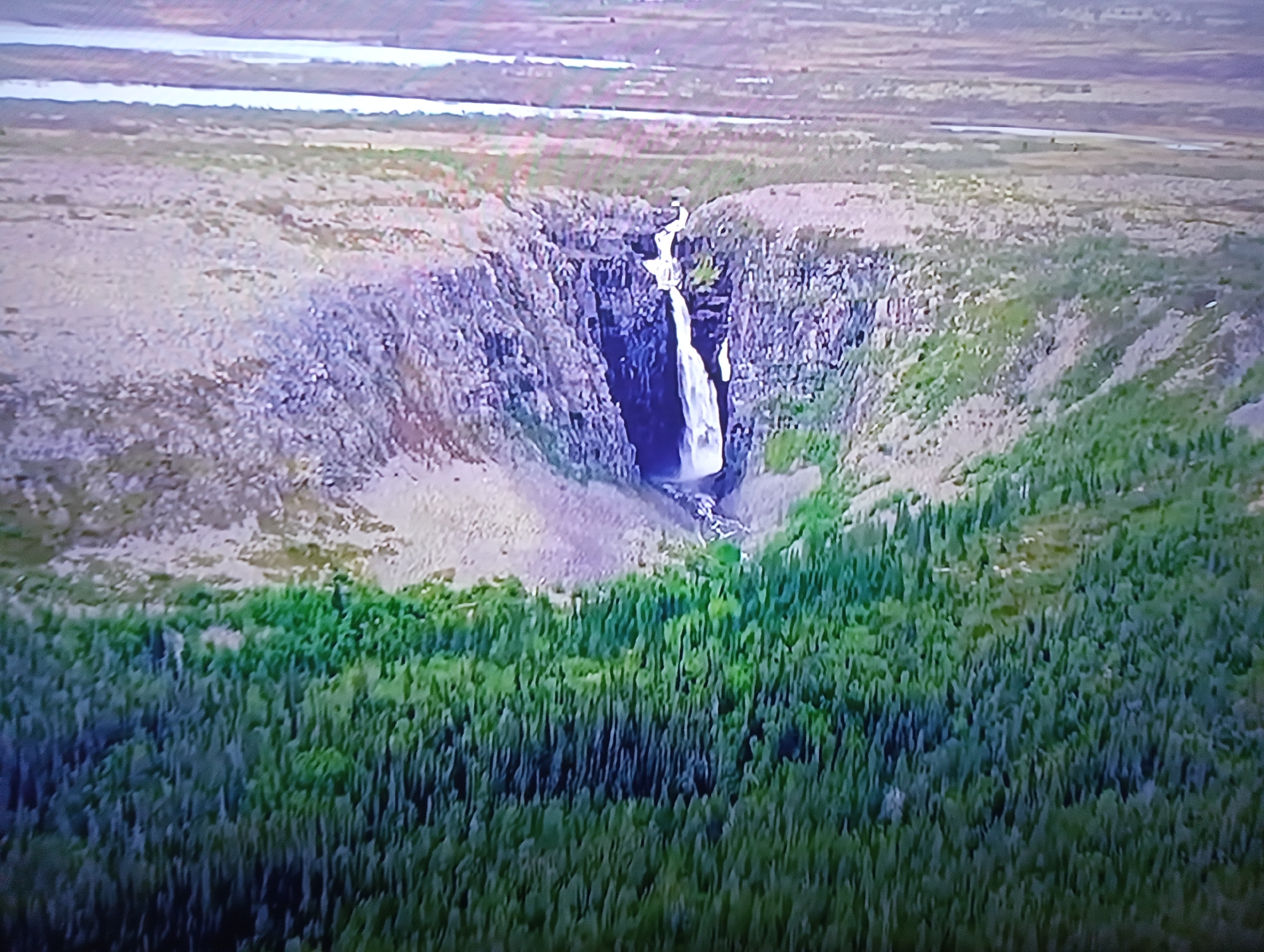 This is an aerial, slightly angled, high-resolution photograph of a waterfall cascading into a narrow canyon. The surrounding landscape consists of barren, rocky terrain and a dense forest of evergreen trees. In the background, a body of water, likely a river or lake, is visible. There are no people or man-made structures in the image, indicating a natural, remote setting. The image does not contain any elements that suggest it is located in the city of Dnipro, Ukraine.