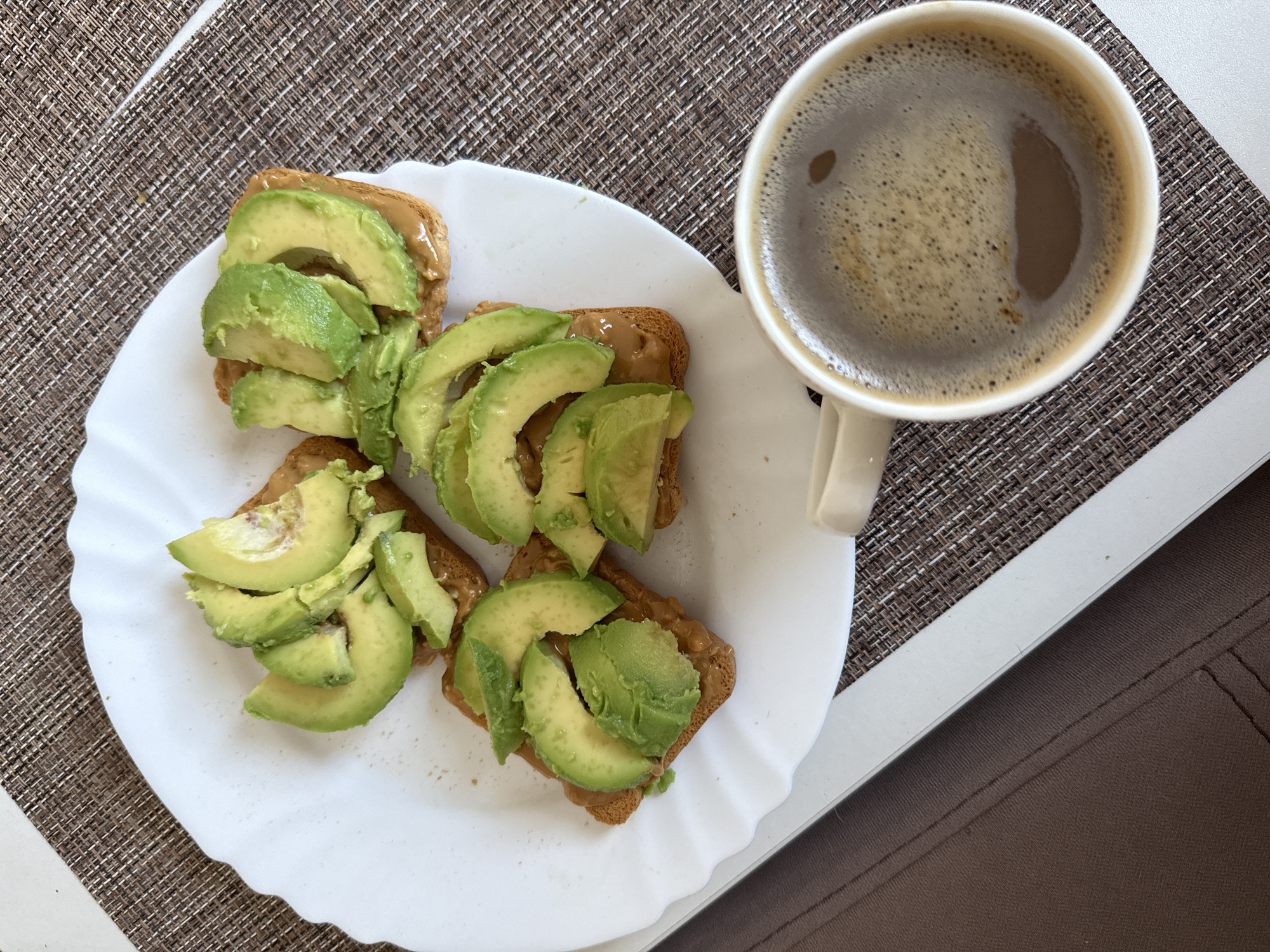 A close-up, overhead shot shows a white, scalloped-edge plate with four pieces of toast topped with peanut butter and sliced avocado. The toast is golden brown and square. The avocado is bright green and sliced thinly. To the right of the plate is a white mug filled with dark coffee, topped with a layer of foam. The plate and mug sit on a textured, brown placemat. The background consists of a brown surface, possibly a table or fabric. The scene is indoors and appears to be a breakfast or snack.