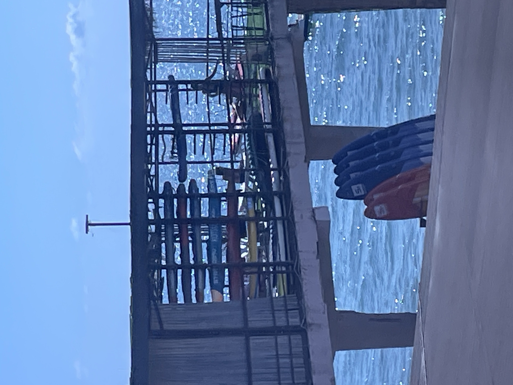 The image depicts a waterfront structure, likely a pier or a boathouse, with a metal rack system holding multiple surfboards. The surfboards are stacked horizontally, with some appearing to be brightly colored like red and yellow, while others are a darker blue. The structure itself is elevated, with supports visible above the water. The water is blue and appears to be choppy, with sunlight reflecting off its surface, creating a sparkling effect. In the background, a clear blue sky with a few wispy clouds is visible. A T-shaped metal pole is also present in the upper portion of the image. The overall scene suggests a location conducive to water sports.