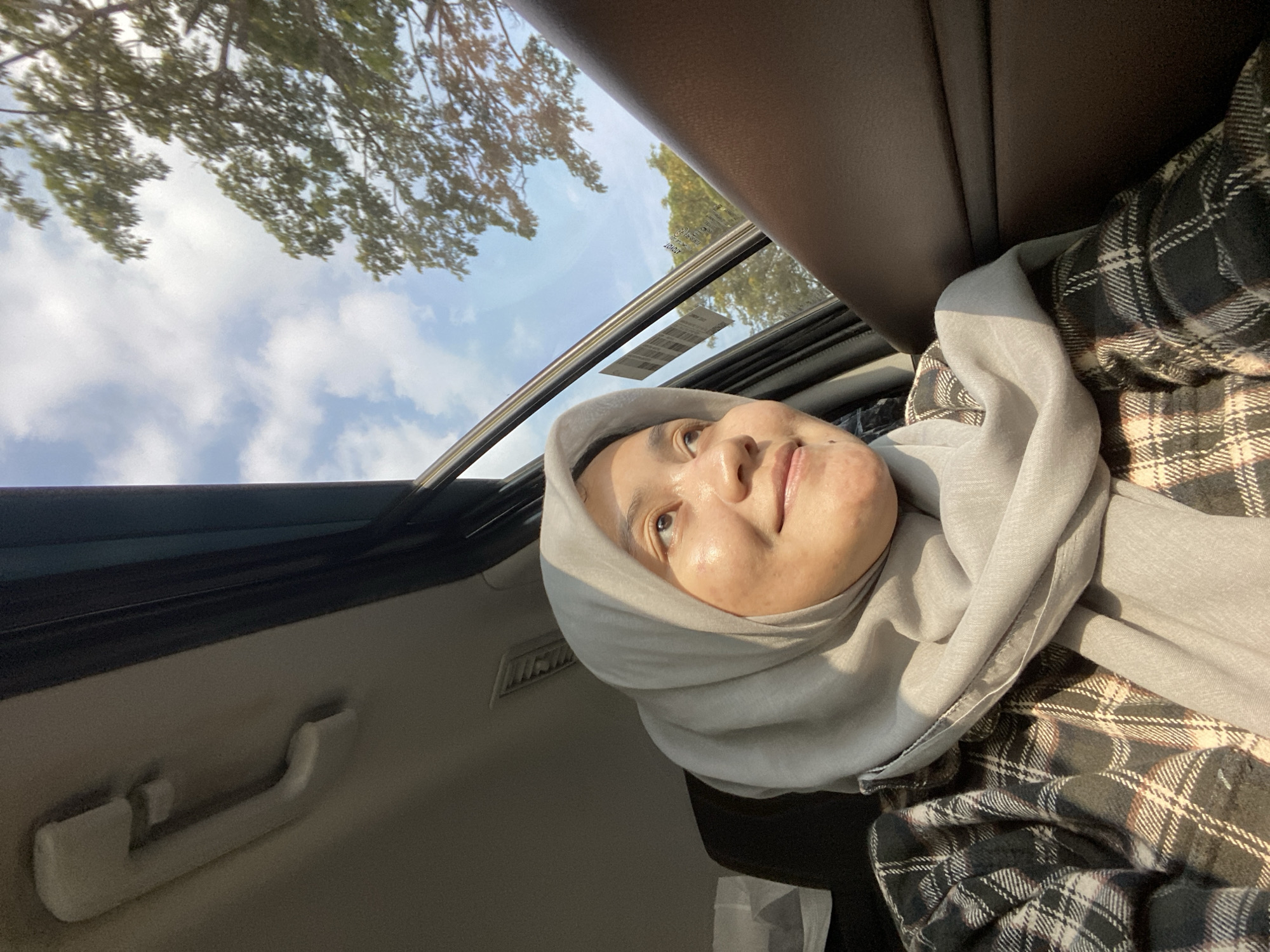 A young woman wearing a light gray hijab and a plaid shirt looks out of a car window. The woman is positioned in the foreground, with her face turned to the side, illuminated by sunlight. Her expression is serene and contemplative. Outside the car, a blue sky with scattered clouds is visible, along with the branches of a leafy tree. The interior of the car, including the door panel and a handle, are also present in the frame. The image was taken in Mandar Sari, Indonesia.