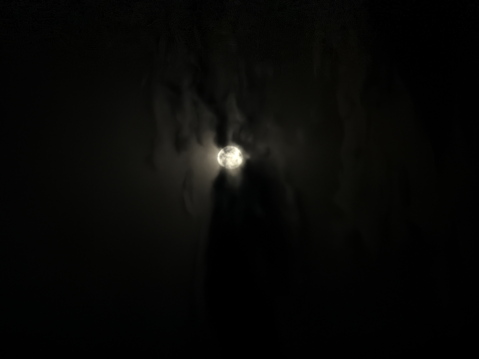 A full moon is visible in the night sky, partially obscured by dark, wispy clouds. The moon is bright and appears to be casting a soft glow on the clouds around it. The overall scene is dark, with the moon being the primary source of light. The image does not contain any recognizable landmarks or features that would indicate a specific location within Lovech, Bulgaria. There are no people or objects depicted in the image.