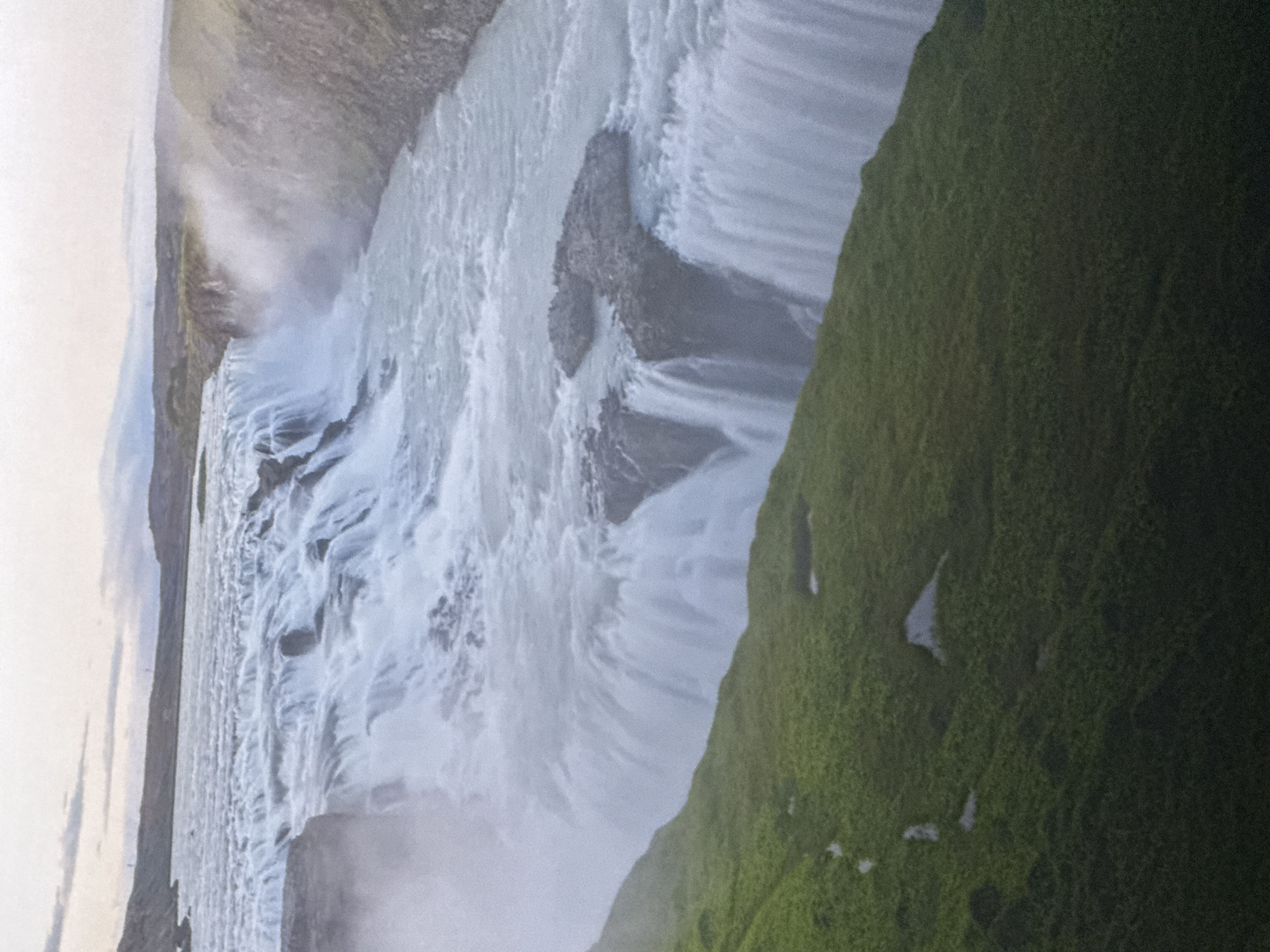 This is an image of a wide, multi-tiered waterfall cascading over rock ledges into a turbulent river below. The water is white and frothy, with a significant amount of mist rising from the falls. The surrounding landscape is characterized by steep, grassy slopes covered in lush green vegetation. In the distance, the sky is overcast with muted, cloudy tones. The scene is a natural landscape, likely a remote or undeveloped area given the lack of any visible man-made structures or human presence. The focus of the image is the powerful movement of the water and the dramatic drop of the waterfall.