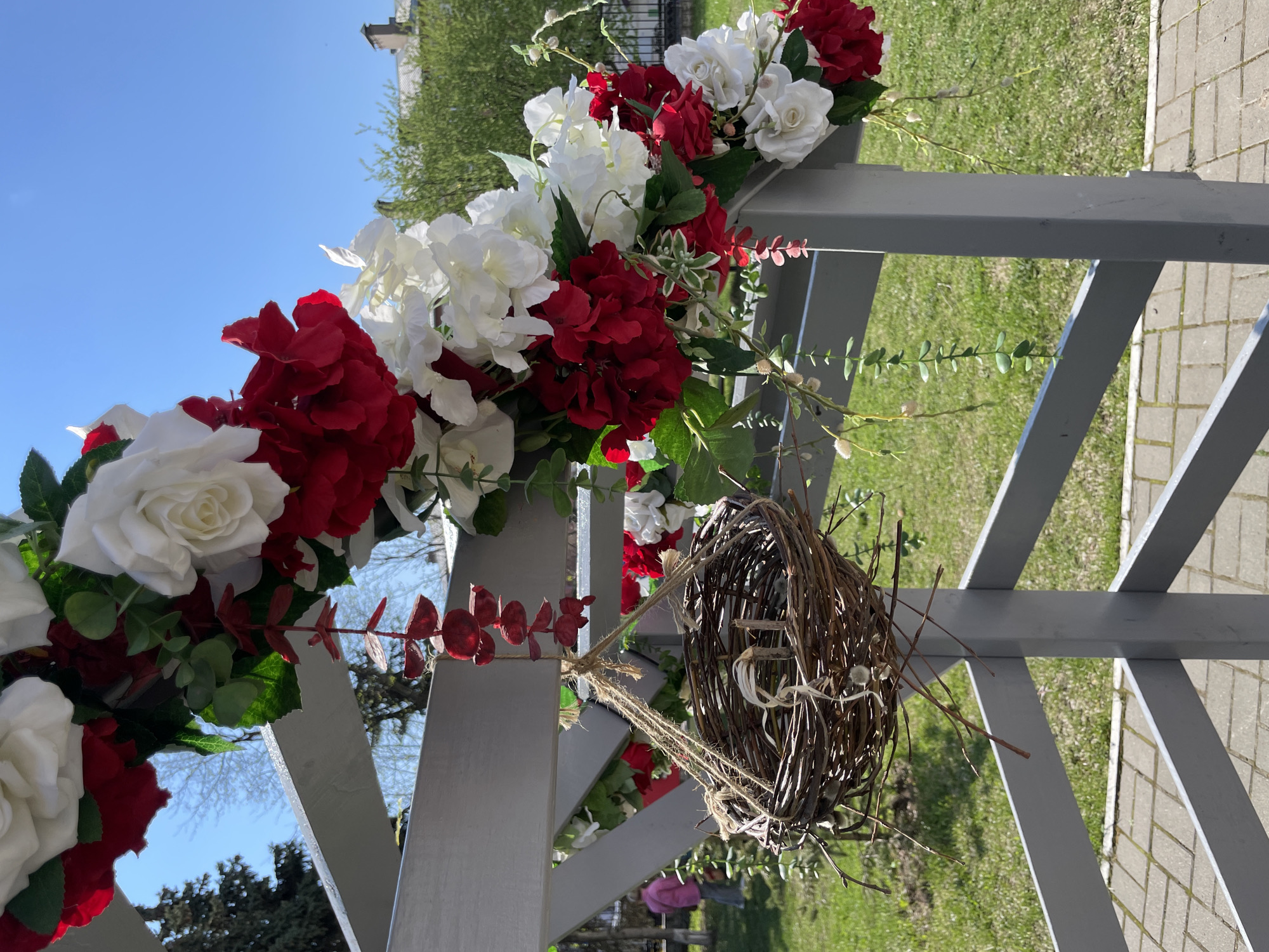 The image shows a white pergola decorated with red and white flowers, possibly hydrangeas and roses. A twig wreath is hanging from the pergola. The background includes a blue sky, trees, grass, and a paved area. No people are readily visible. It is not possible to ascertain whether it is located in the city of Izhevsk, Russia based only on this image.