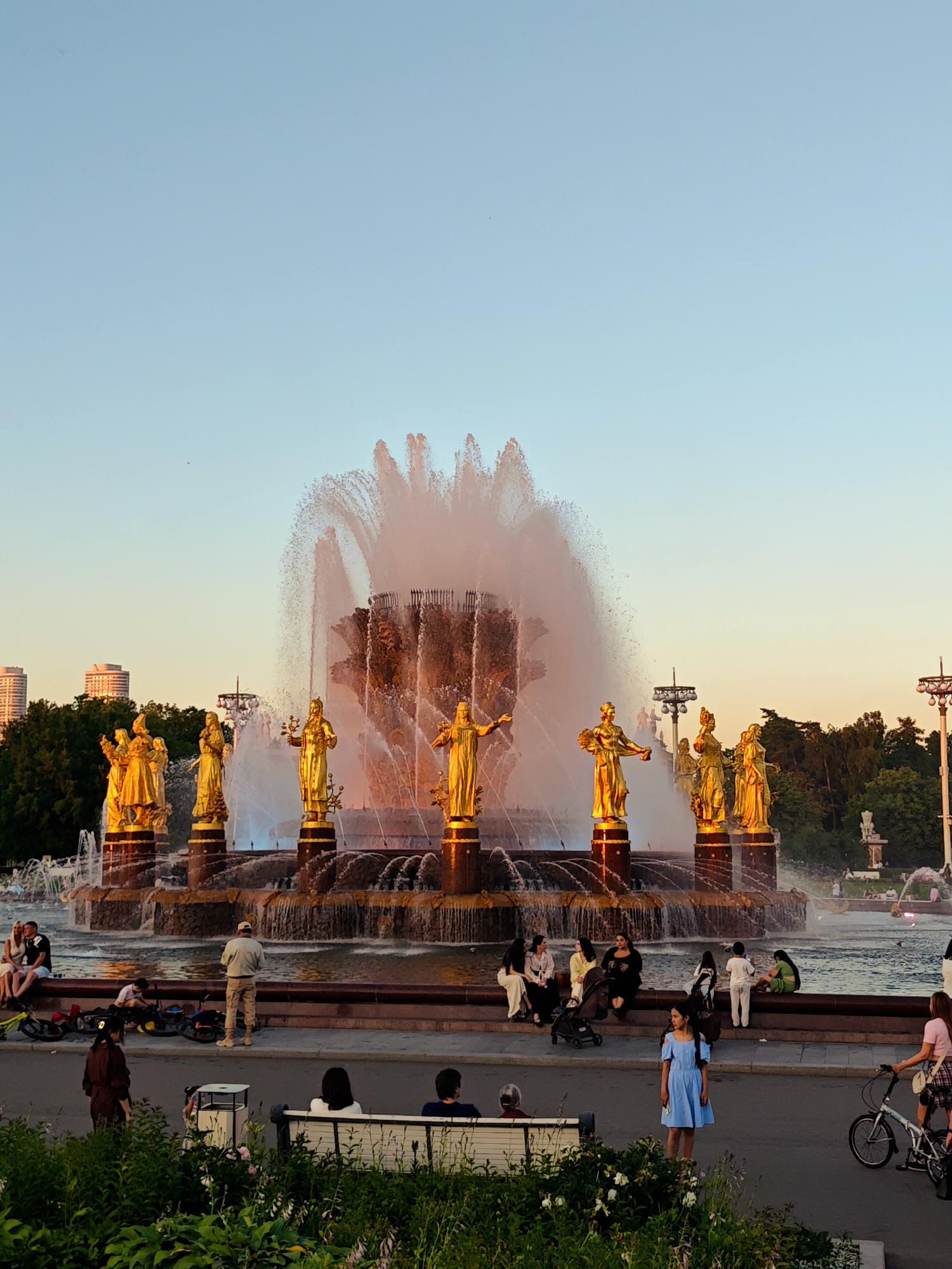 This media file captures a vibrant scene at a large outdoor fountain, likely a tourist attraction in Moscow, Russia. The fountain is the central subject, featuring numerous golden statues of women, possibly allegorical figures, adorned with various items. Water sprays dramatically from the fountain's core, creating a shimmering cascade that is illuminated by the warm glow of the setting sun.
In the foreground and surrounding the fountain, several people are visible, suggesting a leisurely gathering. Some are seated on benches or the fountain's edge, while others stand or walk. The presence of a stroller and bicycles indicates a family-friendly atmosphere. A young girl in a blue dress stands out, looking towards the viewer, while a woman with a bicycle rides past. In the background, tall buildings and lush green trees suggest an urban park or public space.
The time of day is clearly late afternoon or early evening, indicated by the soft, warm light and the clear, pale blue sky. The overall mood appears relaxed and pleasant, with people enjoying the outdoor space and the impressive fountain. There is no visible text in the image.